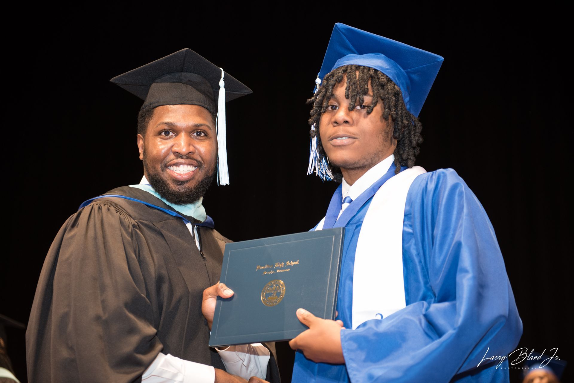 Man in brown graduation gown presents diploma to person in blue graduation gown.