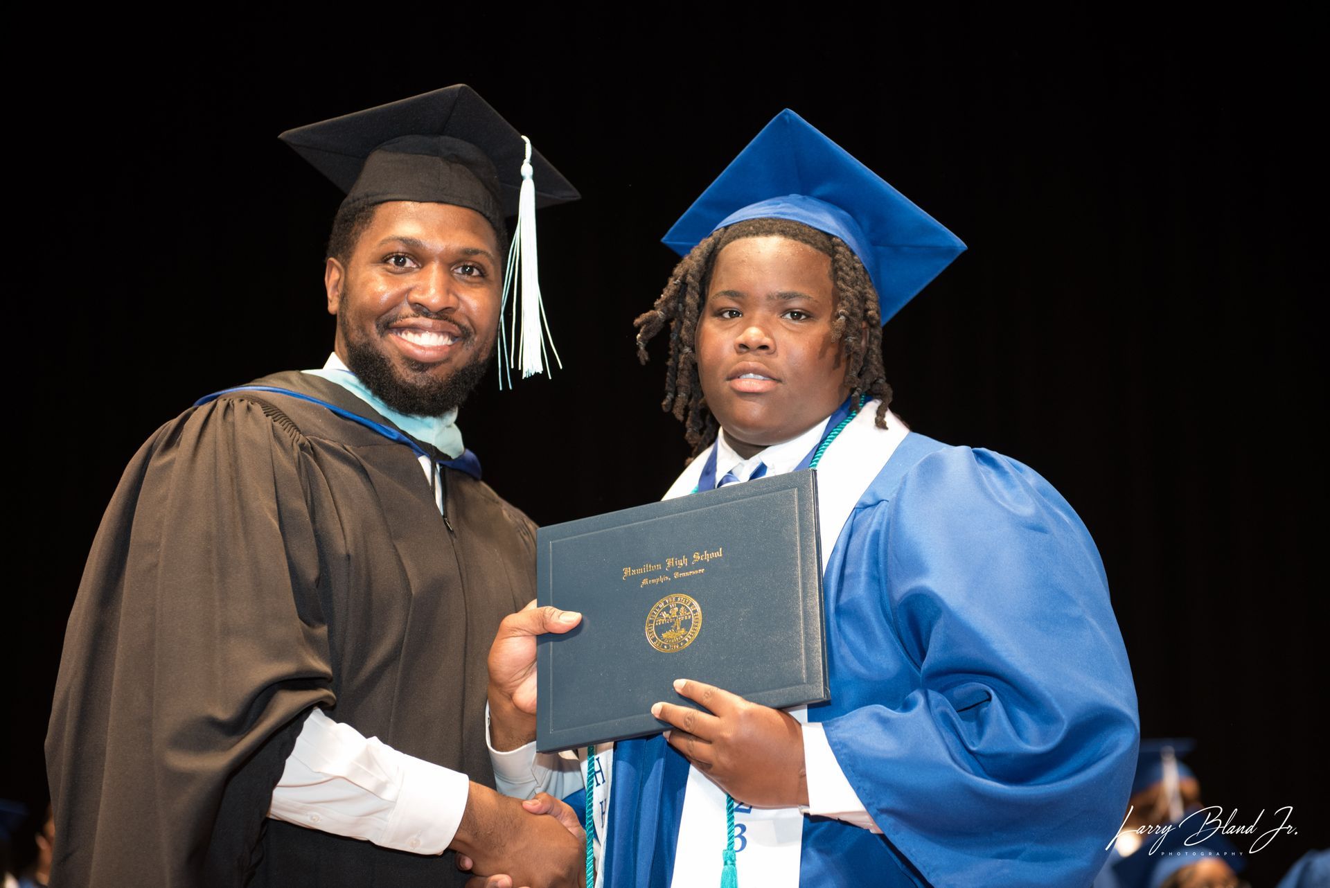 A person in a graduation gown presents a diploma to a student in a blue gown at a ceremony.