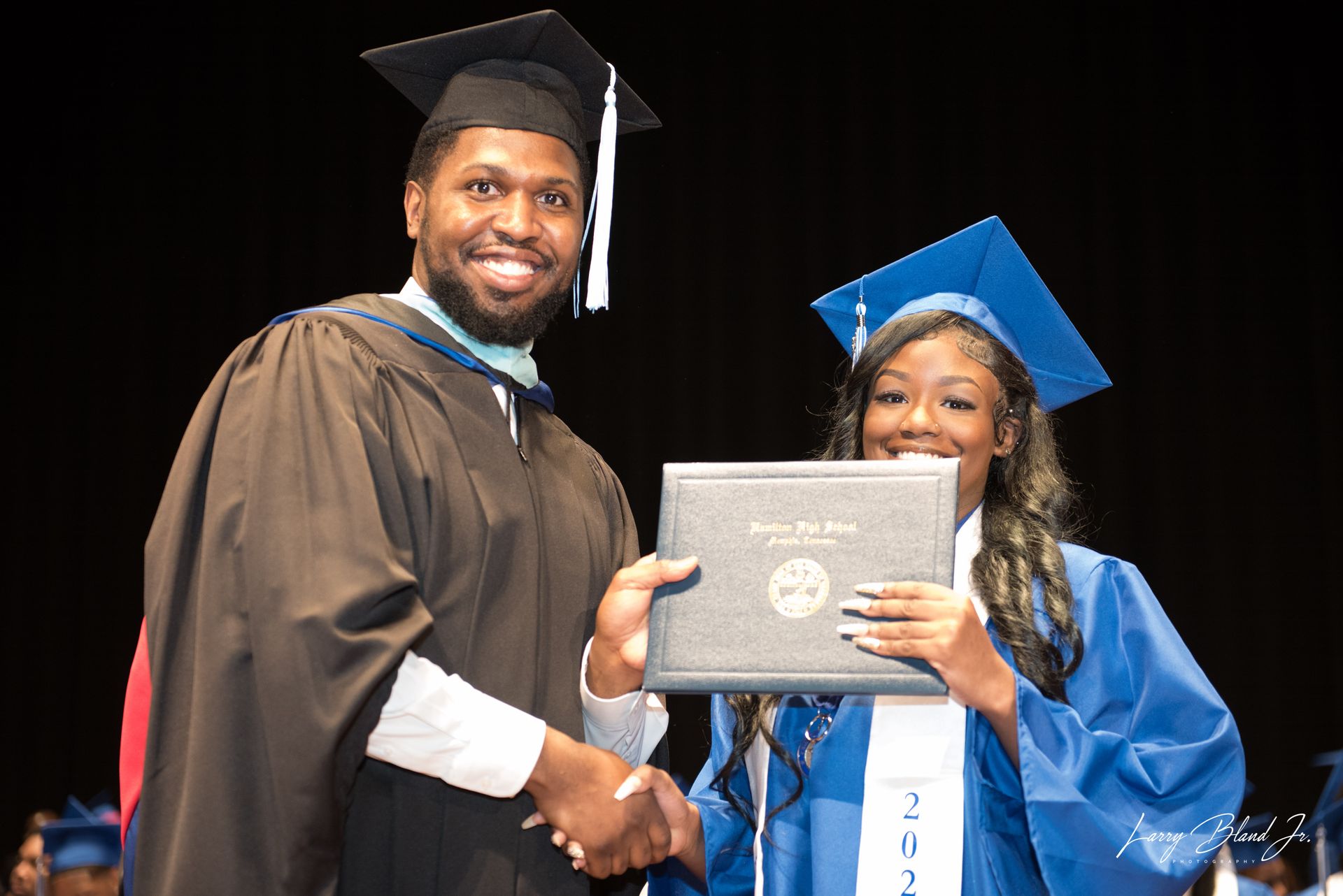Person in cap and gown presenting diploma to graduate, both smiling at ceremony.