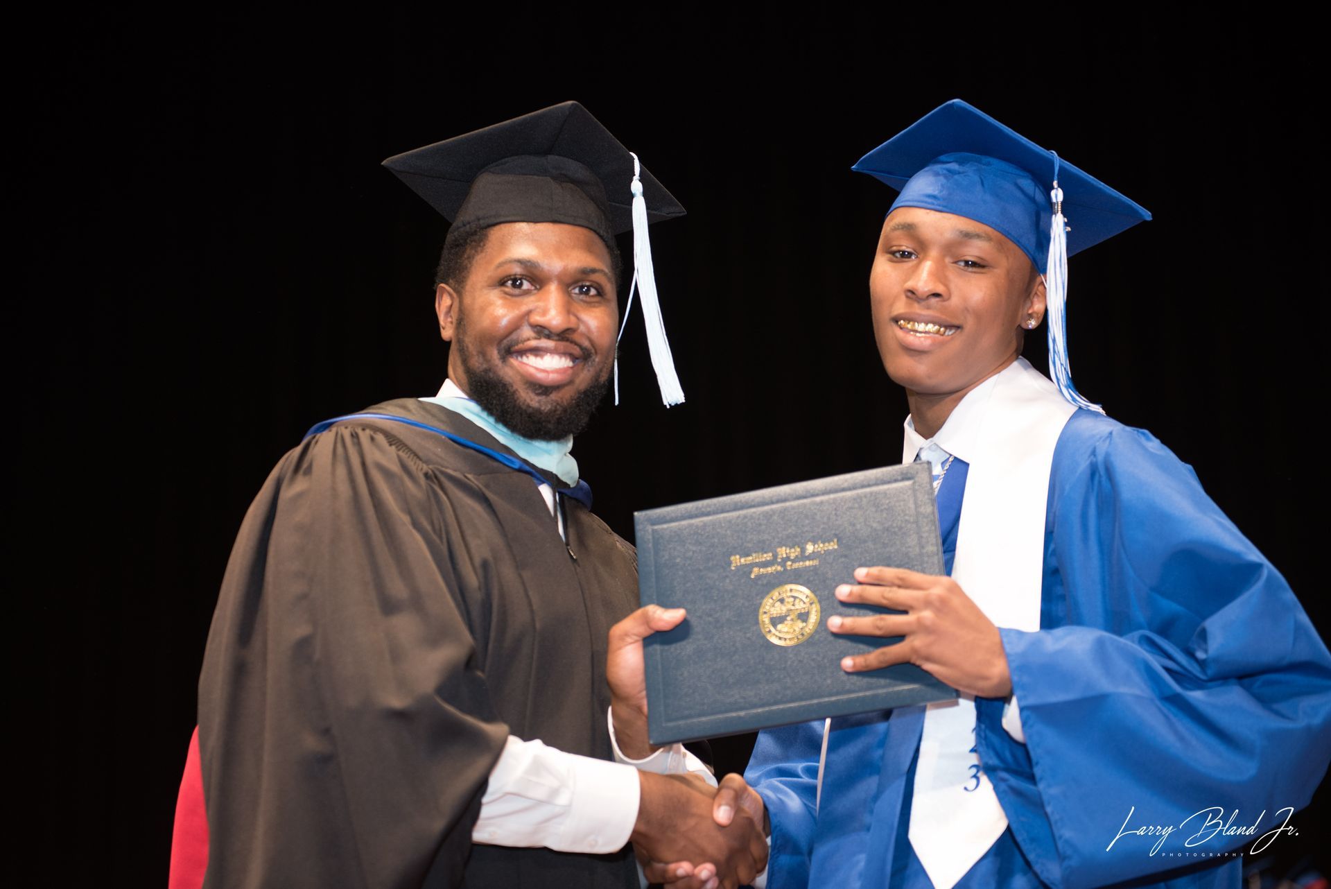 Man in cap and gown handing diploma to a graduate in a blue gown, shaking hands, stage backdrop.