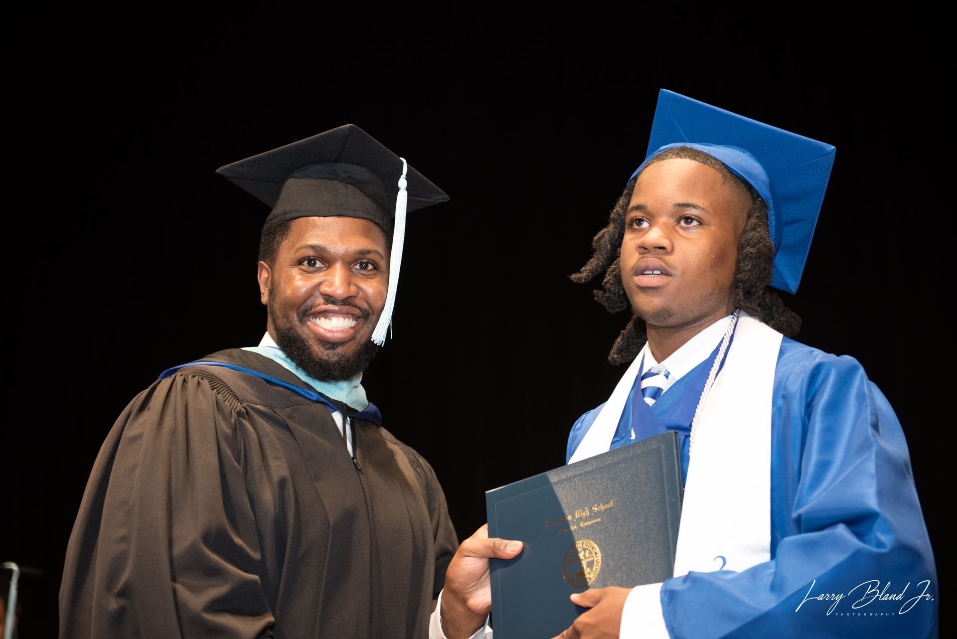 Man in graduation gown hands diploma to person in blue cap and gown.