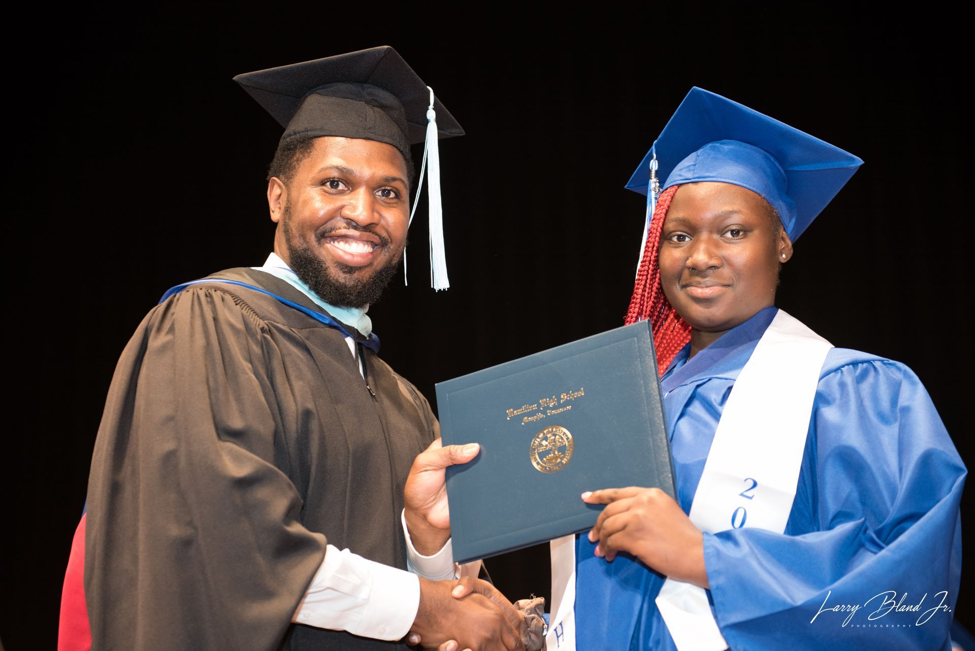 A graduate in blue robe receiving a diploma from a person in a black gown during a ceremony.
