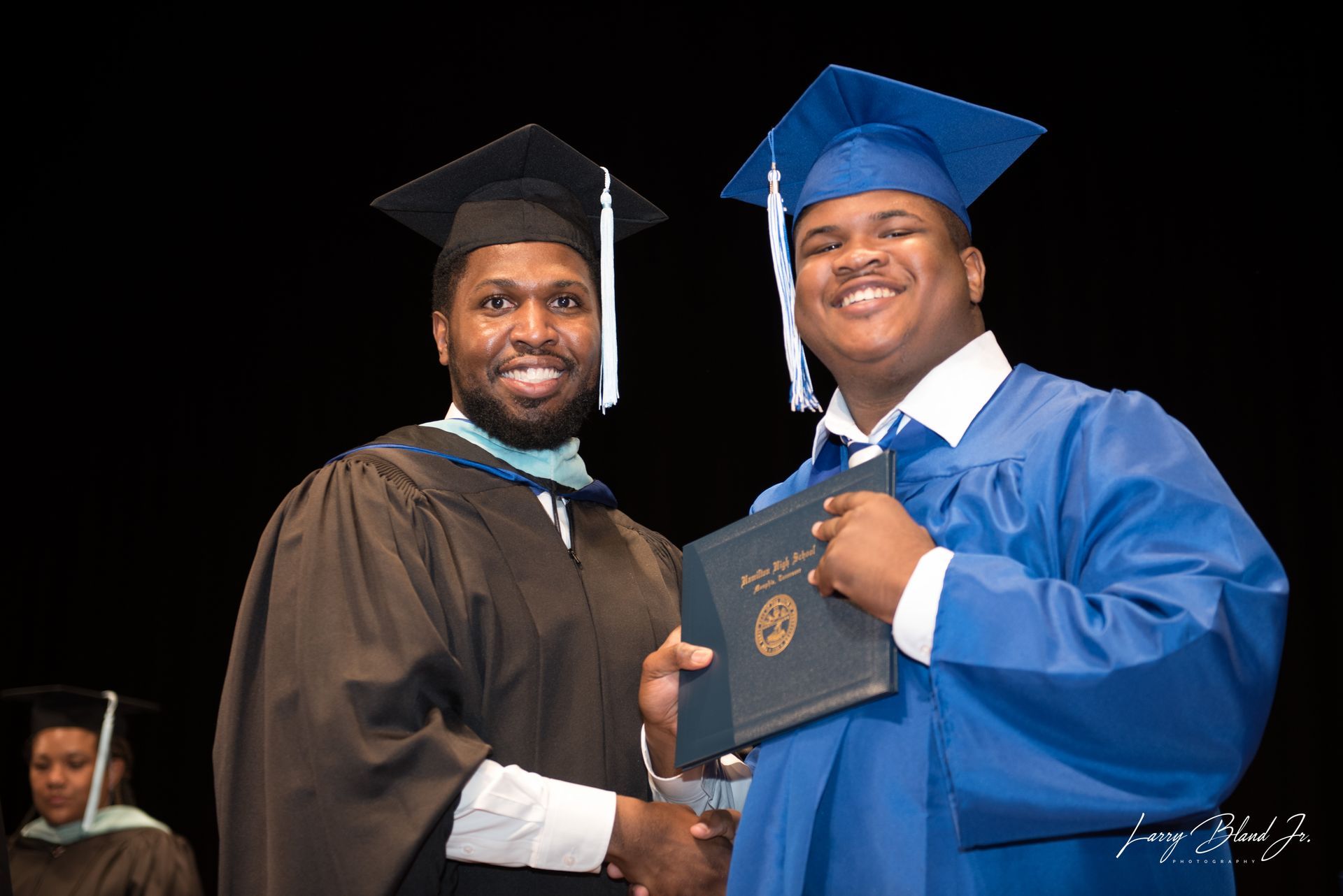 Man in black robe hands diploma to a smiling person in a blue graduation gown.