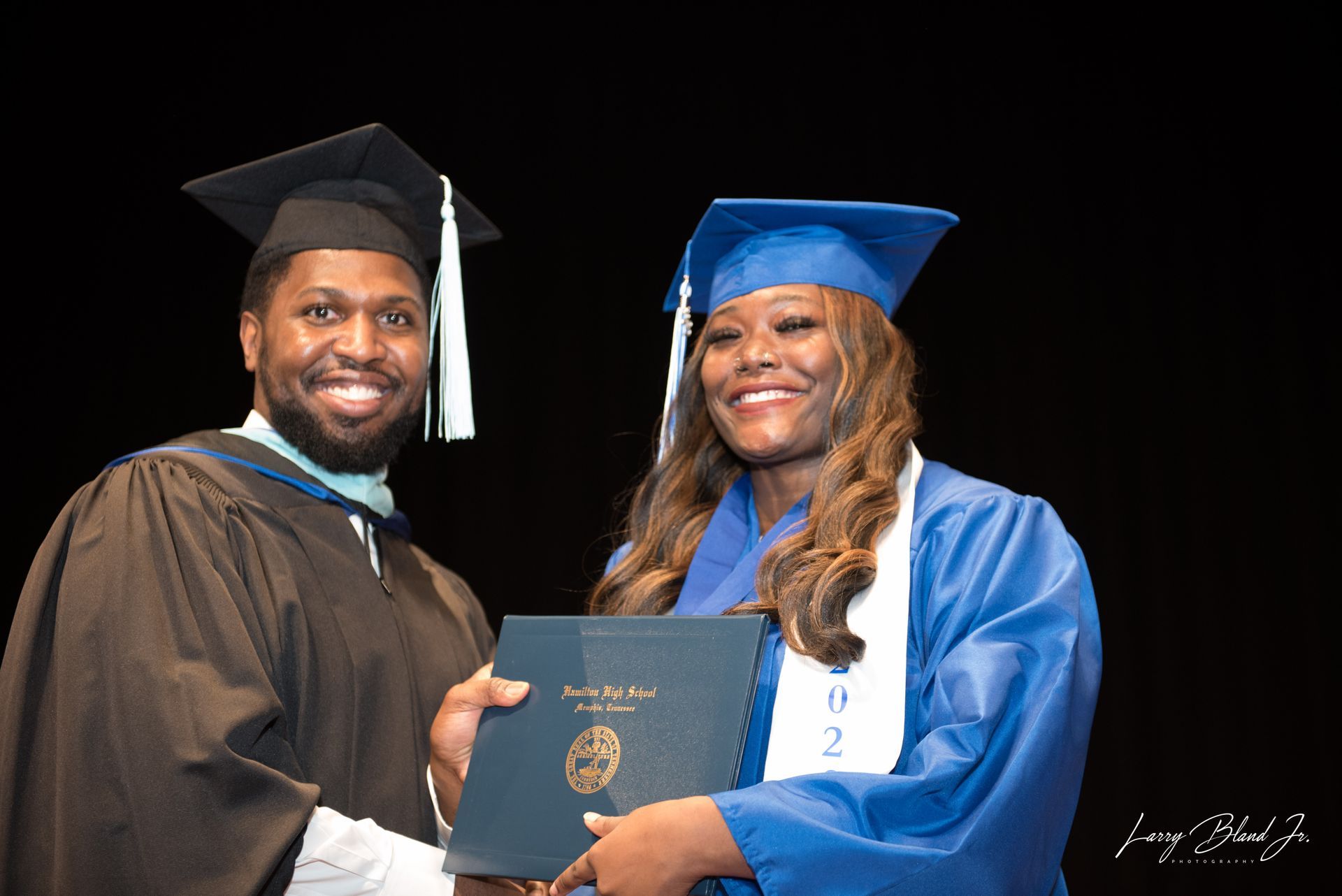 Person in cap and gown presents diploma to a smiling graduate in blue gown. Stage backdrop.