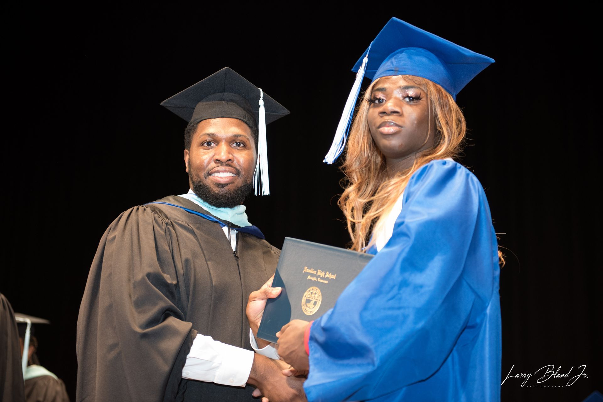 A graduate in a blue gown receiving a diploma from a person in academic regalia on a stage.