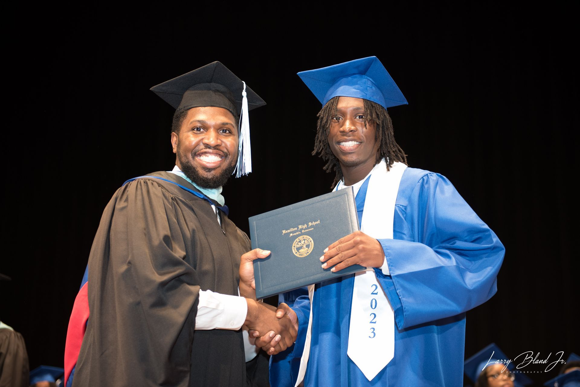 A graduate in blue robe shaking hands with a robed man, receiving diploma on stage. 2023.