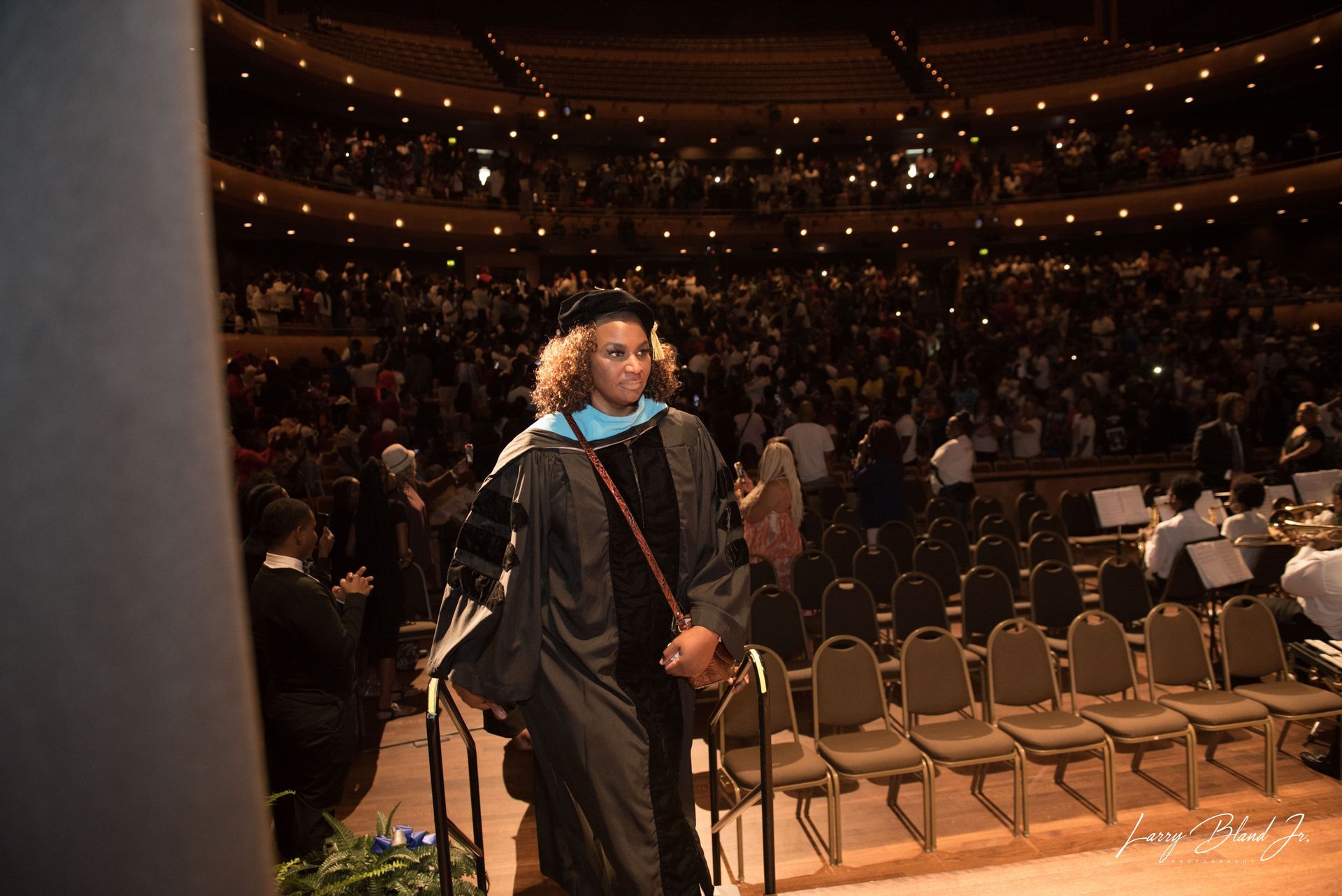 Person in graduation regalia walks on stage, auditorium crowd in background.