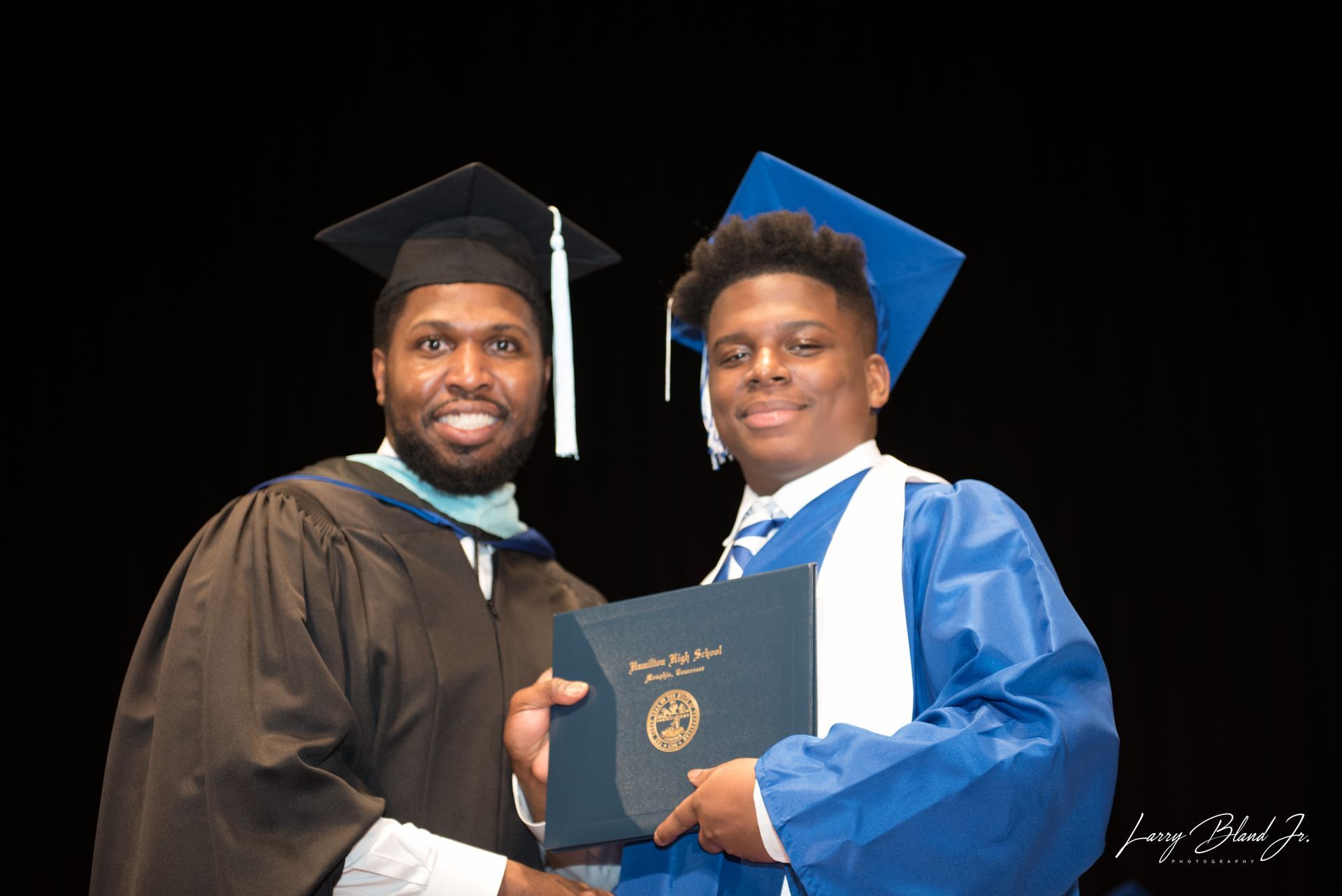 Man in brown robe hands a diploma to a person in a blue graduation gown on a black background.