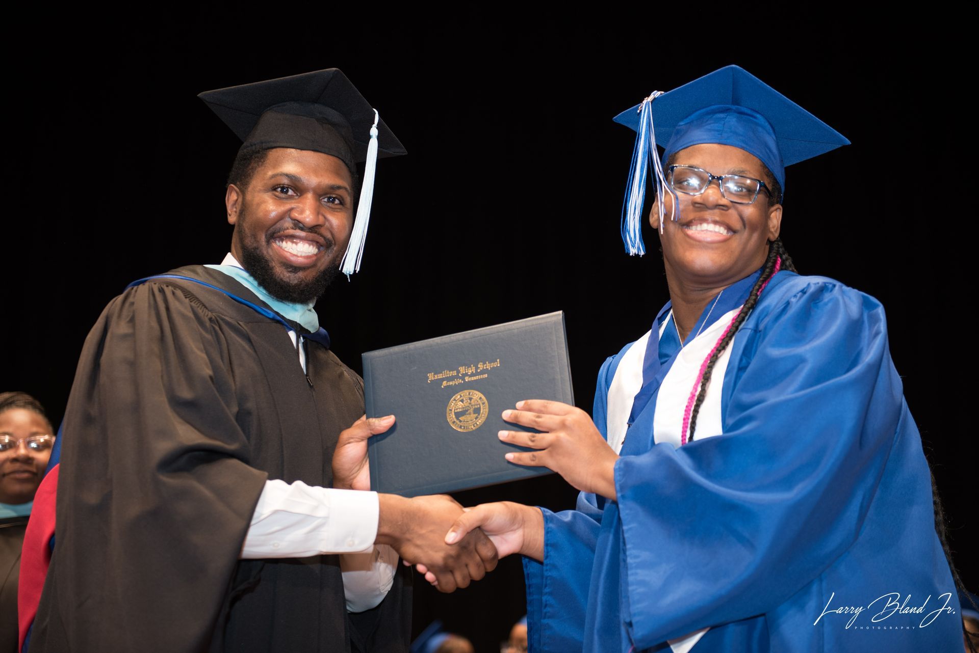 Man in cap and gown handing diploma to person in graduation attire on stage.