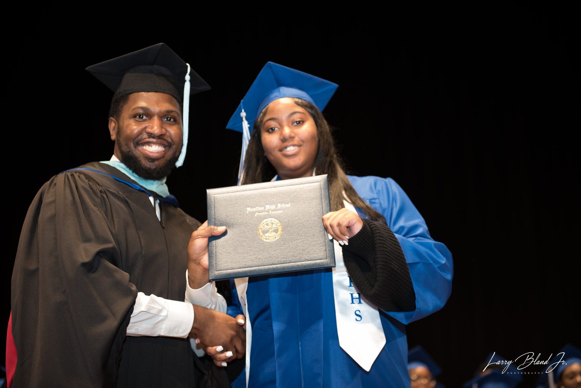 A person in a black gown hands a diploma to someone in a blue graduation gown on stage.