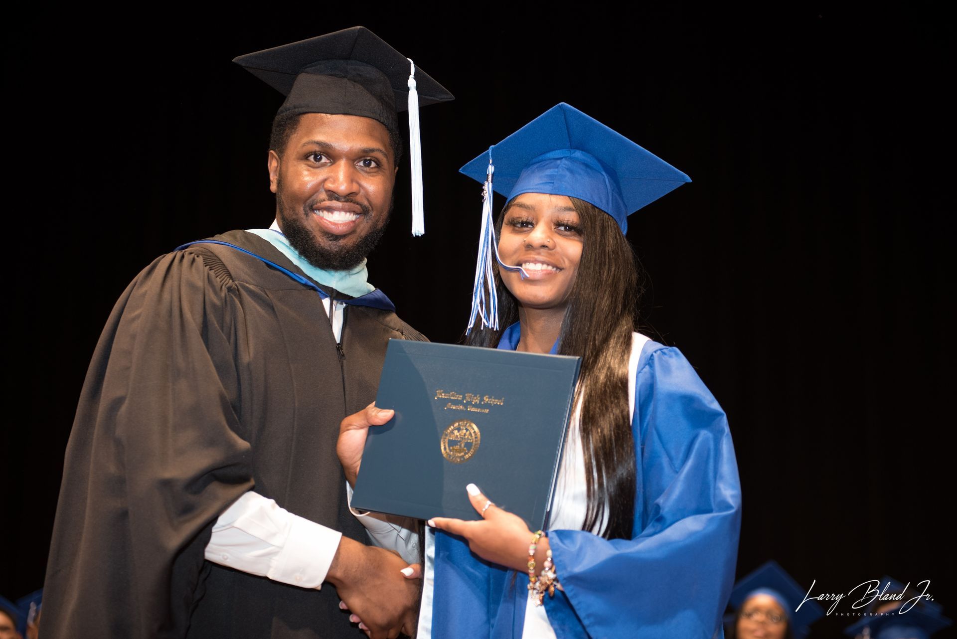 Graduation: Man in gown hands diploma to person in blue cap and gown on stage.