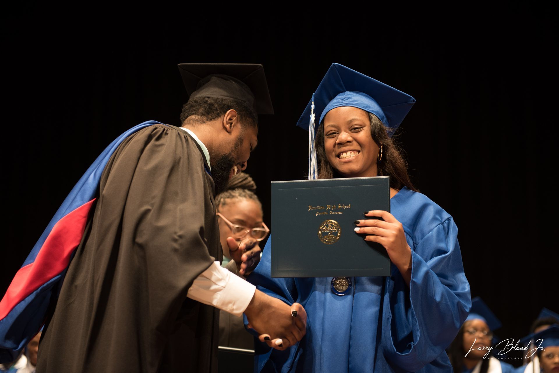 Person in blue cap and gown receives diploma during graduation, shaking hands with a person in academic attire.