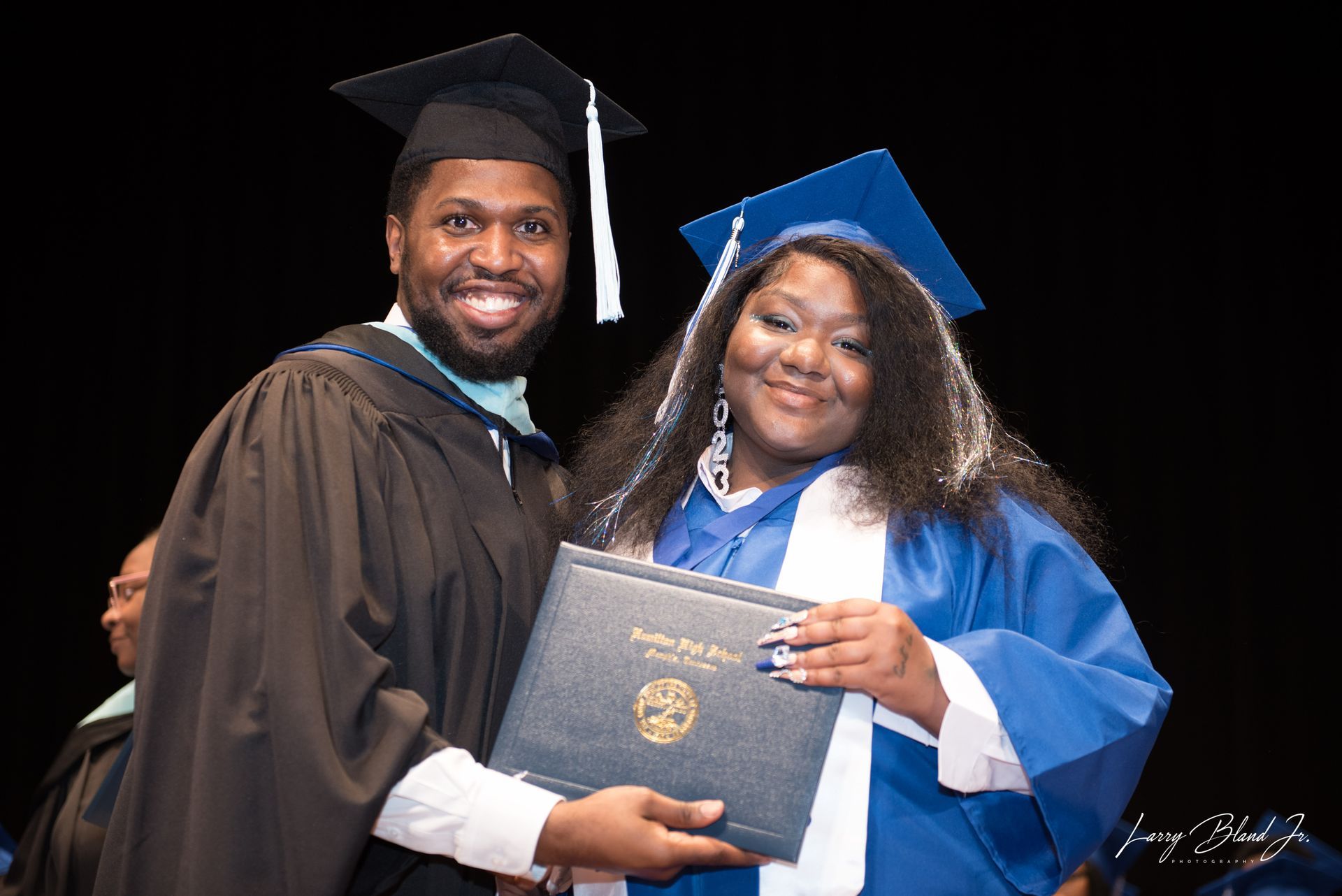 Person in cap and gown presenting diploma to graduate in blue gown at ceremony. Both smiling.