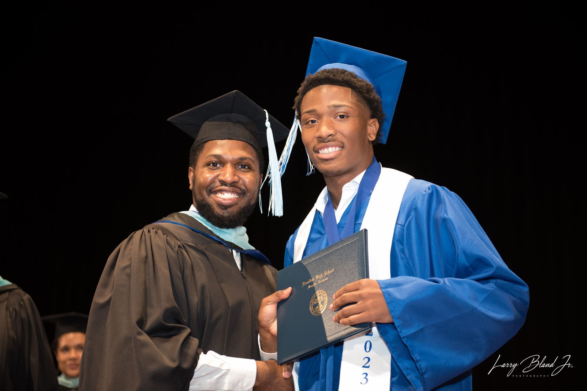 Two people at a graduation ceremony. One presents a diploma to the other, both in caps and gowns.