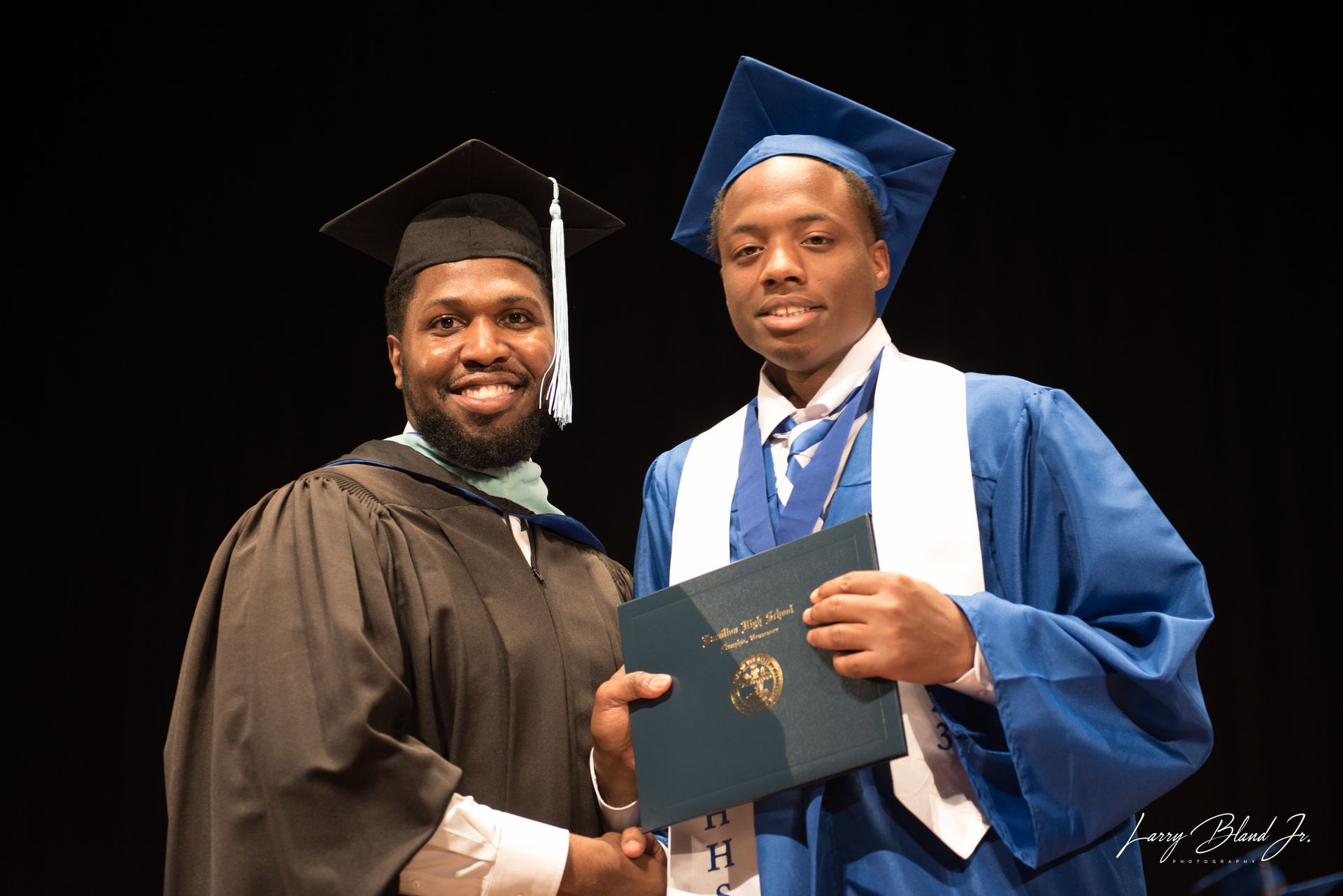 Man in graduation gown hands a diploma to a person wearing a blue cap and gown on stage.
