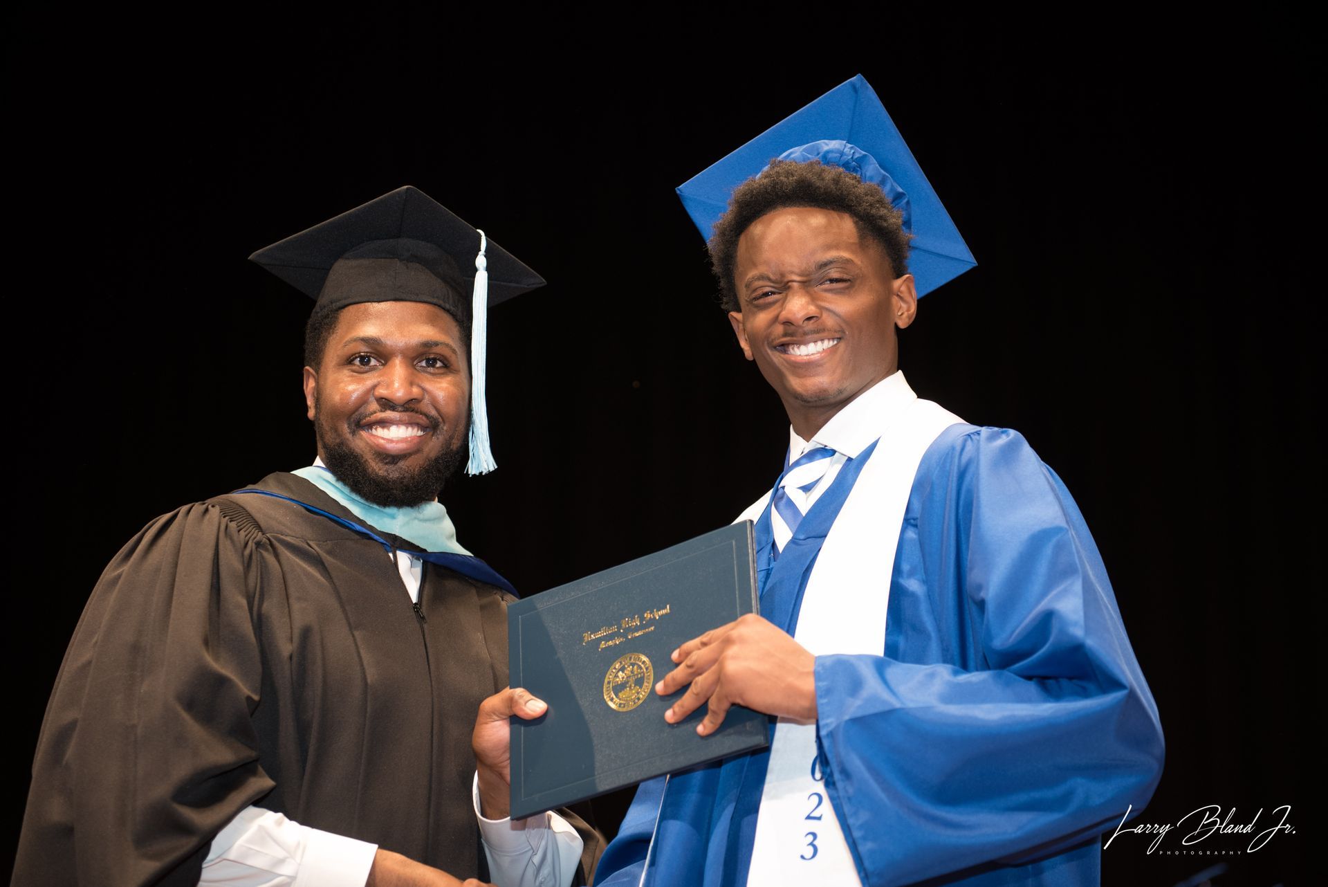Two men at a graduation ceremony. One in cap and gown presents diploma to graduate in blue gown, both smiling.
