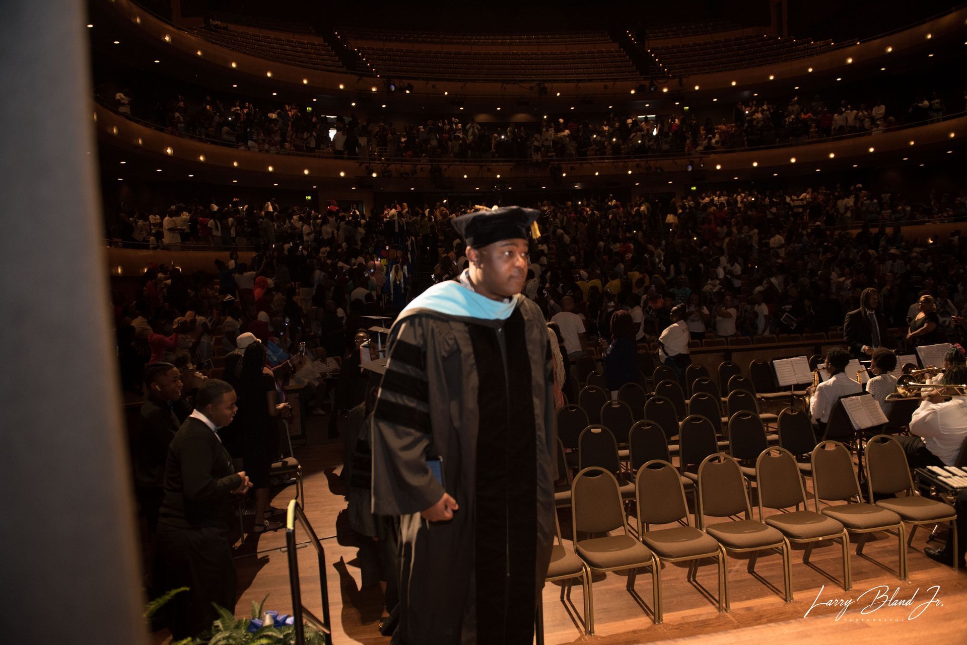 Person in graduation gown on stage, audience in background.
