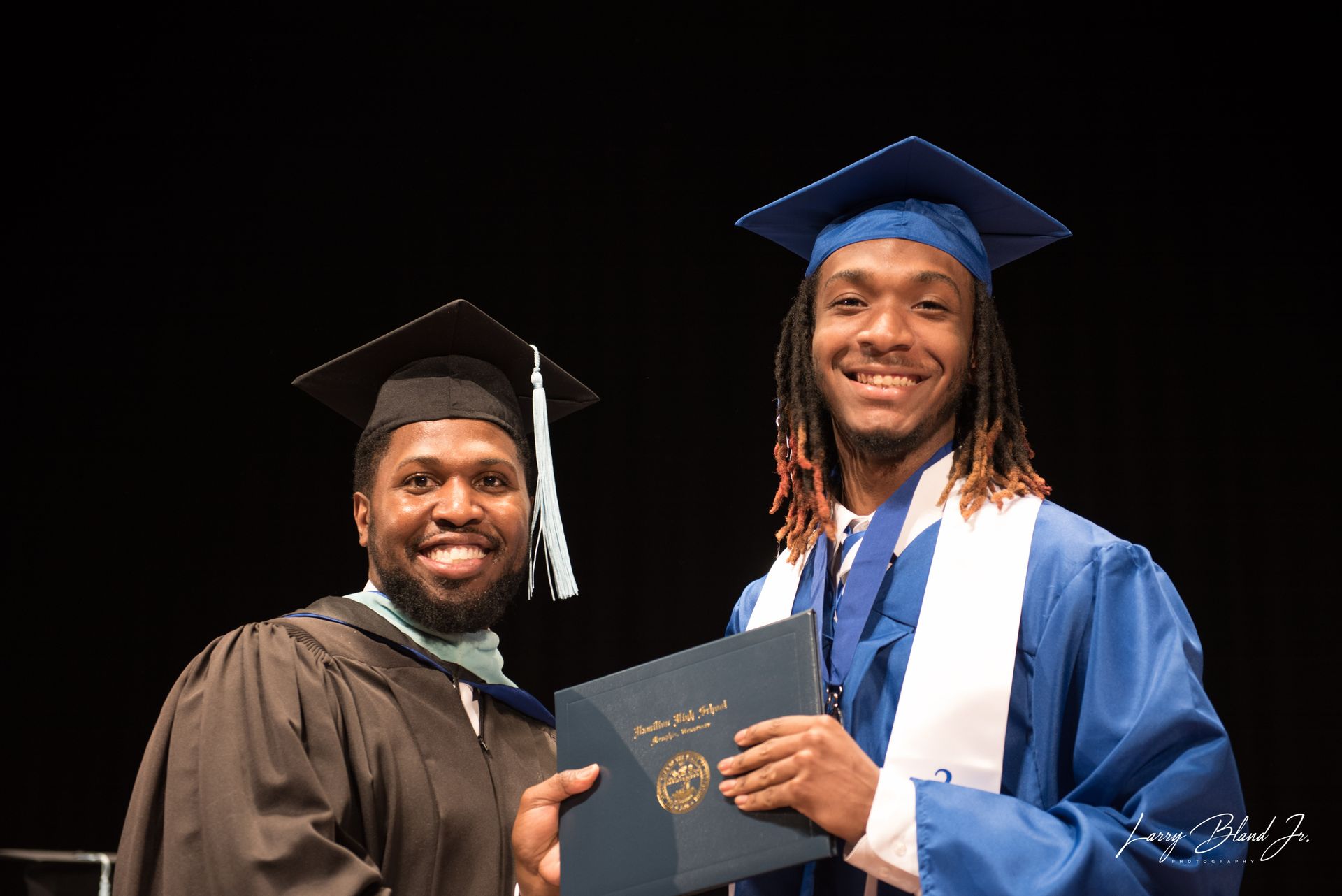 Man in blue graduation gown receives diploma from man in black gown. Both smiling.