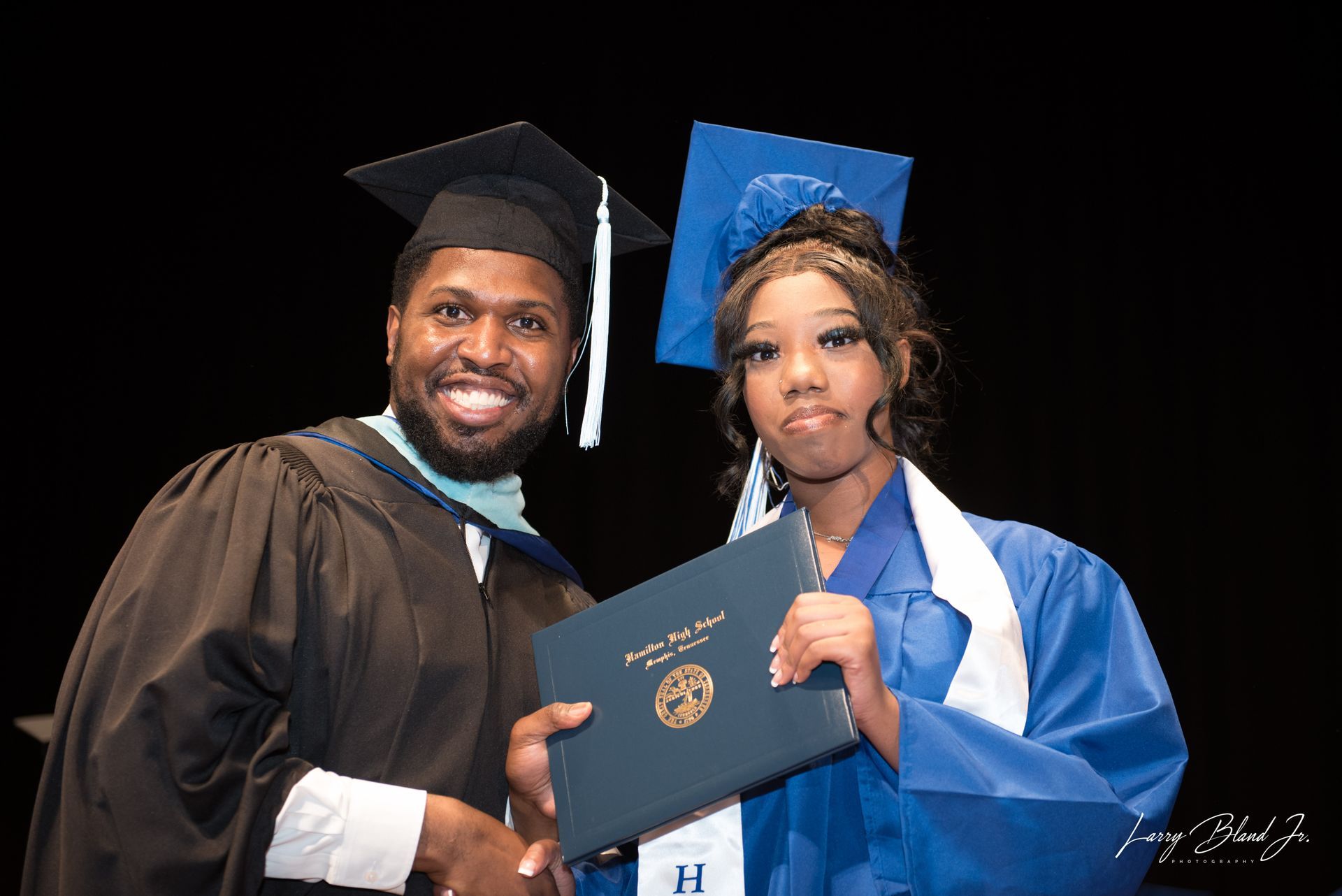 Man in graduation gown hands diploma to person in blue gown and cap.