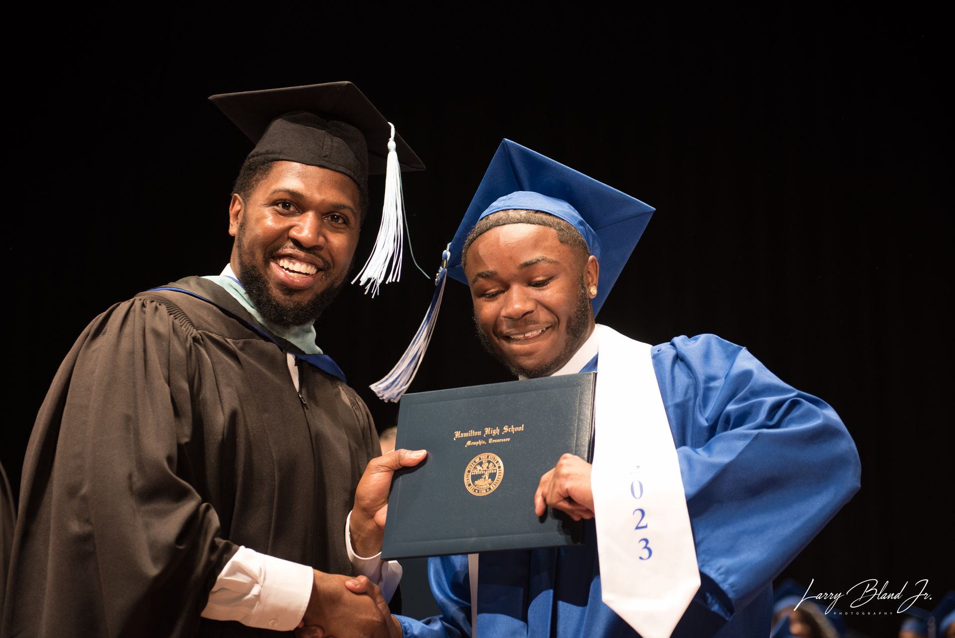 Man in black graduation gown hands diploma to person in blue gown, both smiling.