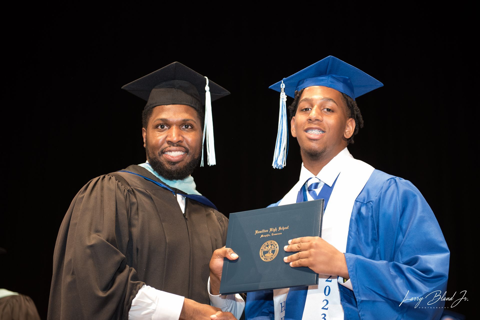 Man in black gown hands diploma to person in blue graduation attire.