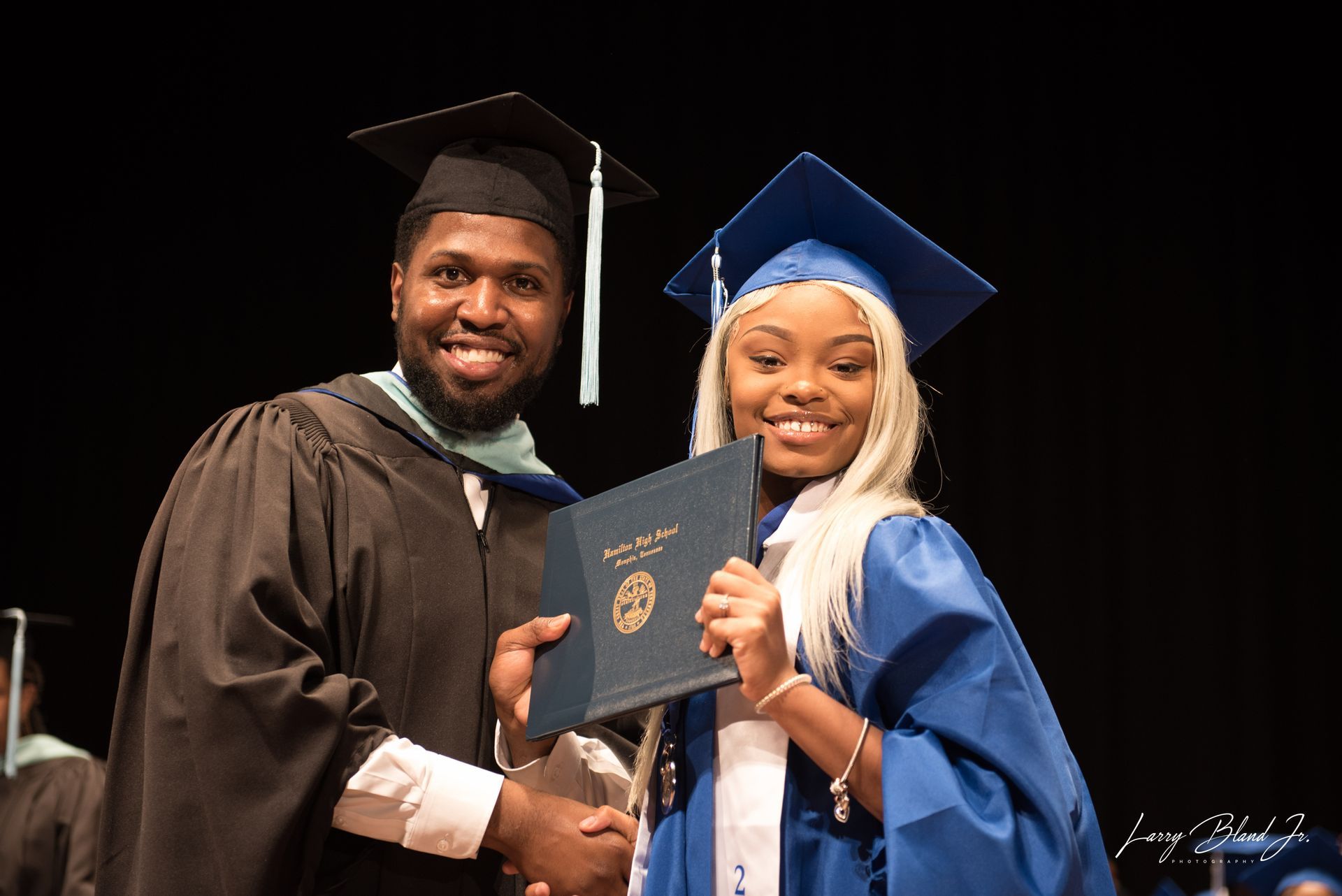 Man in black robe hands diploma to person in blue graduation gown. Smiling, stage setting.