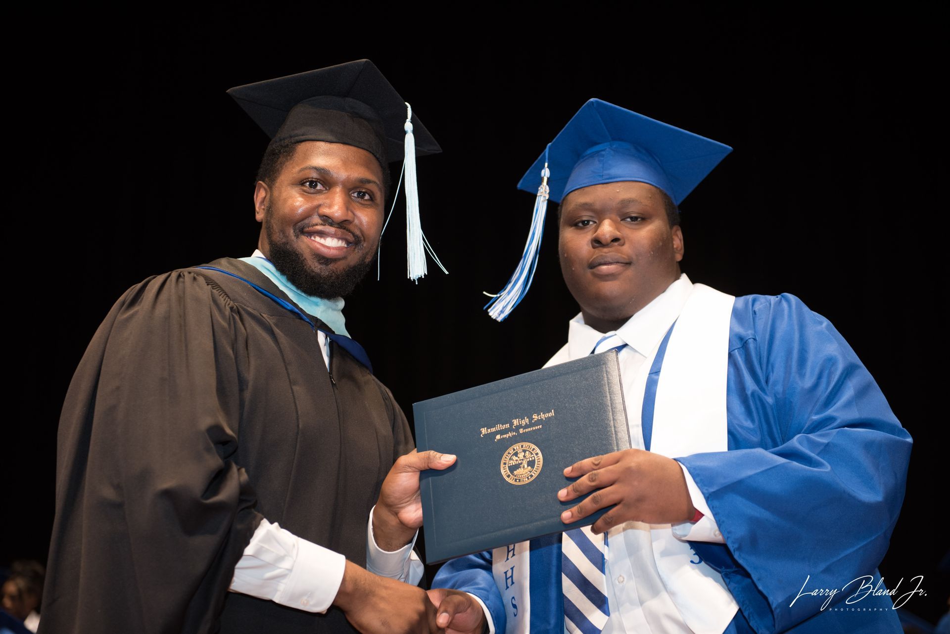 Man in black graduation attire hands diploma to a graduate in blue attire.