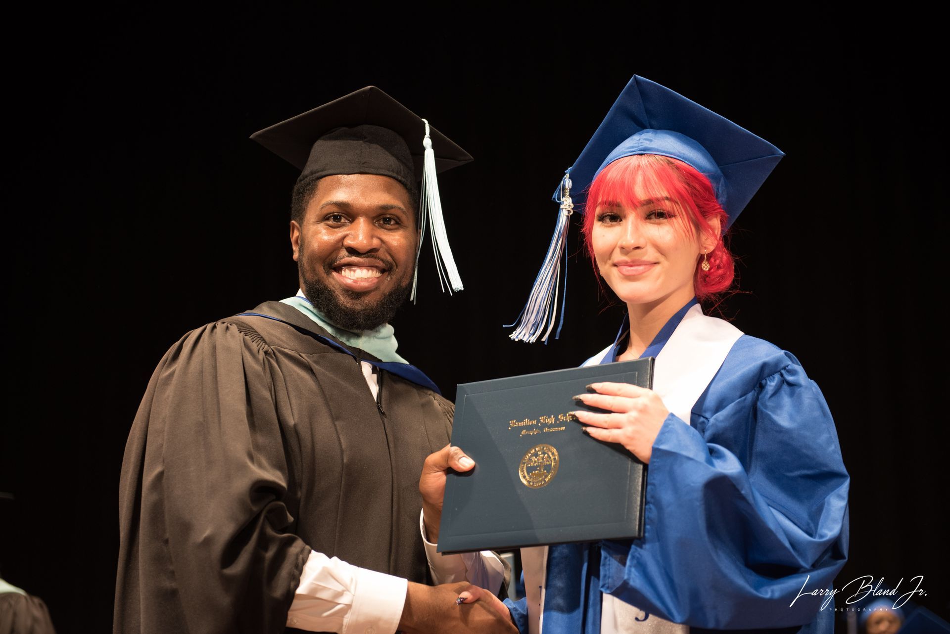 Person in black graduation attire hands diploma to person in blue attire onstage.