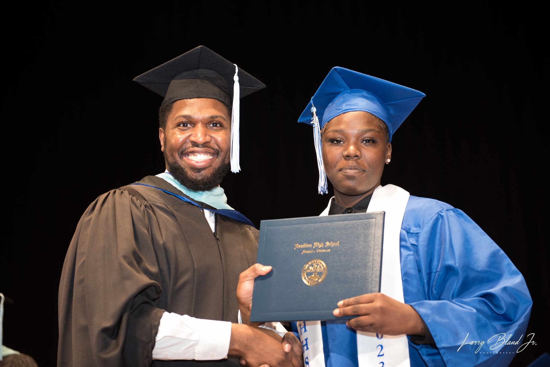 Person in black gown handing a diploma to a person in blue graduation gown, both smiling. Stage setting.