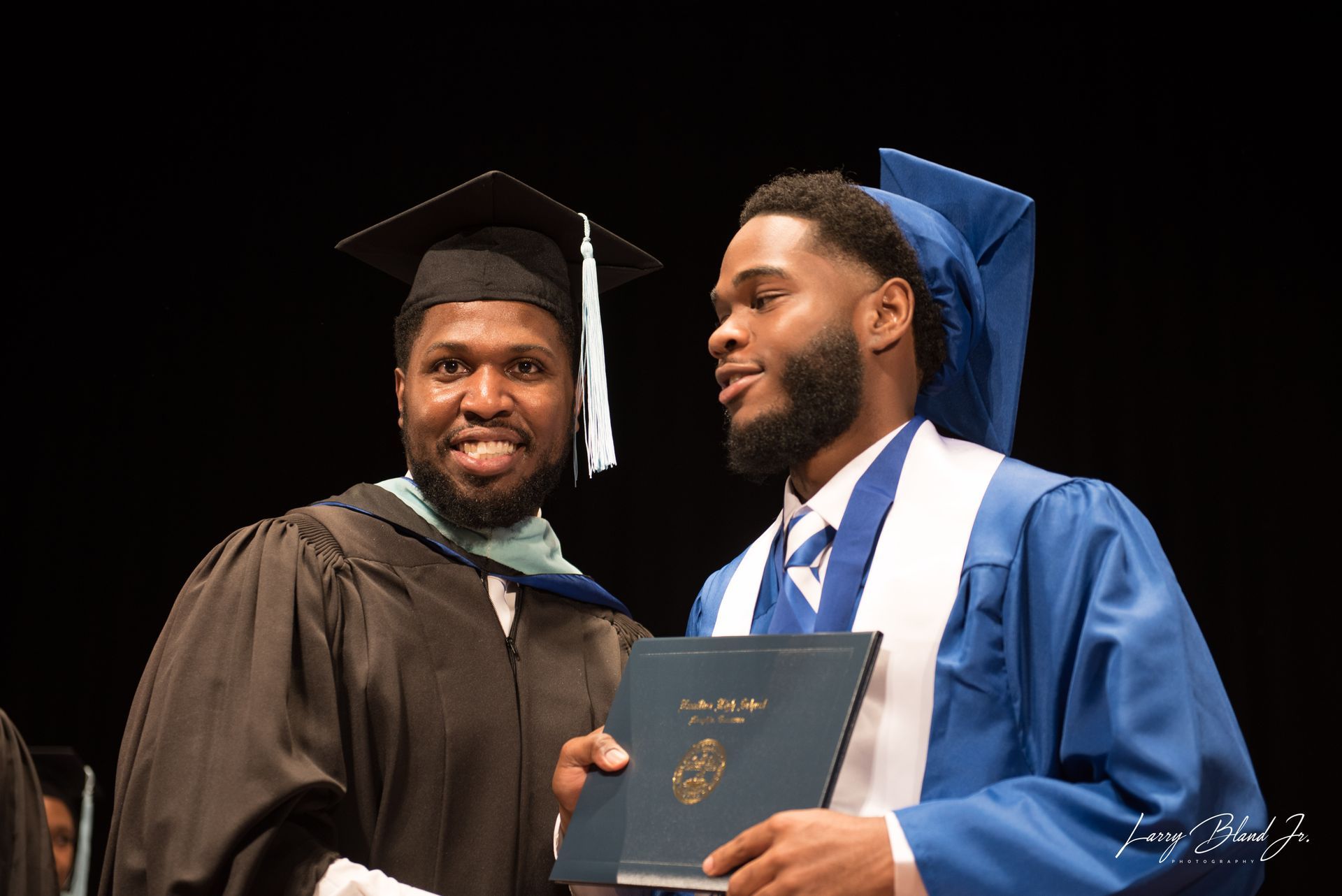 Man in black gown hands diploma to man in blue graduation robe.