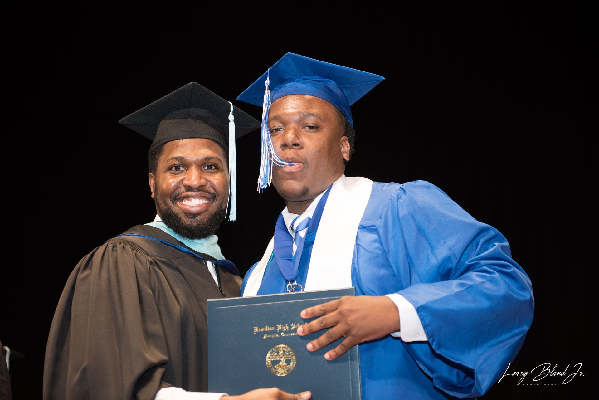 Two men in graduation attire pose with a diploma, one in blue gown, the other in black; smiling.