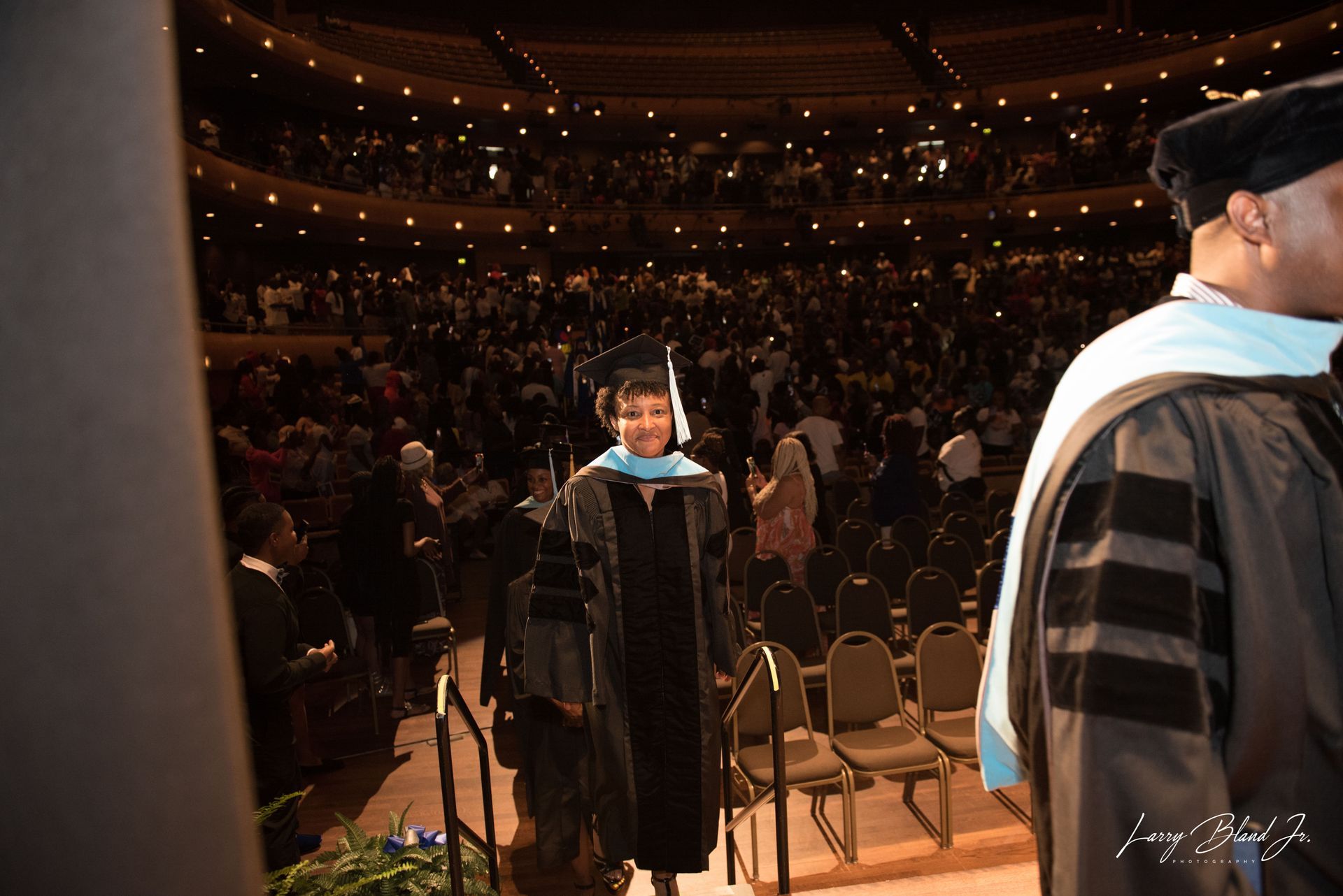Graduation ceremony. Person in cap and gown walks toward stage, crowd in background.