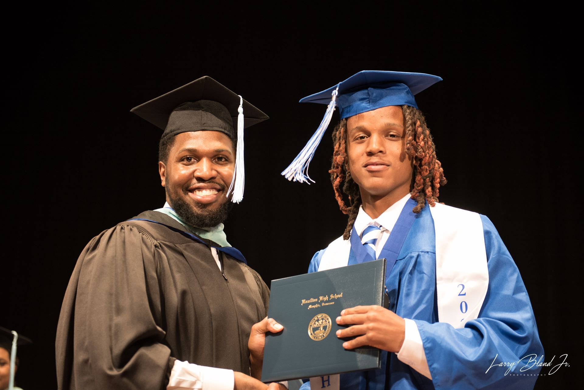 Man in graduation gown handing diploma to a graduate wearing a blue cap and gown.