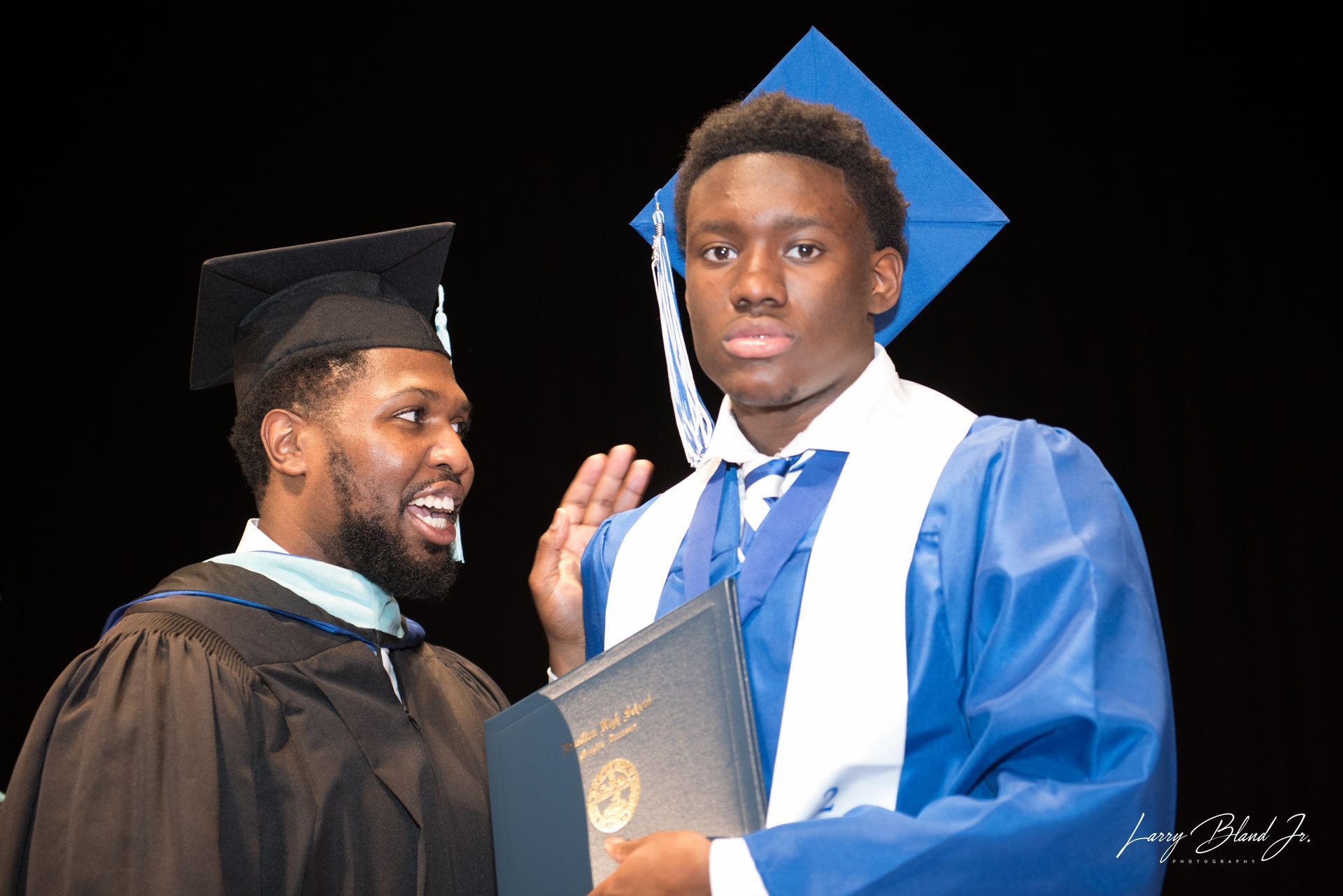 Man in black graduation gown congratulates another in blue, holding a diploma, with a blue square cap.