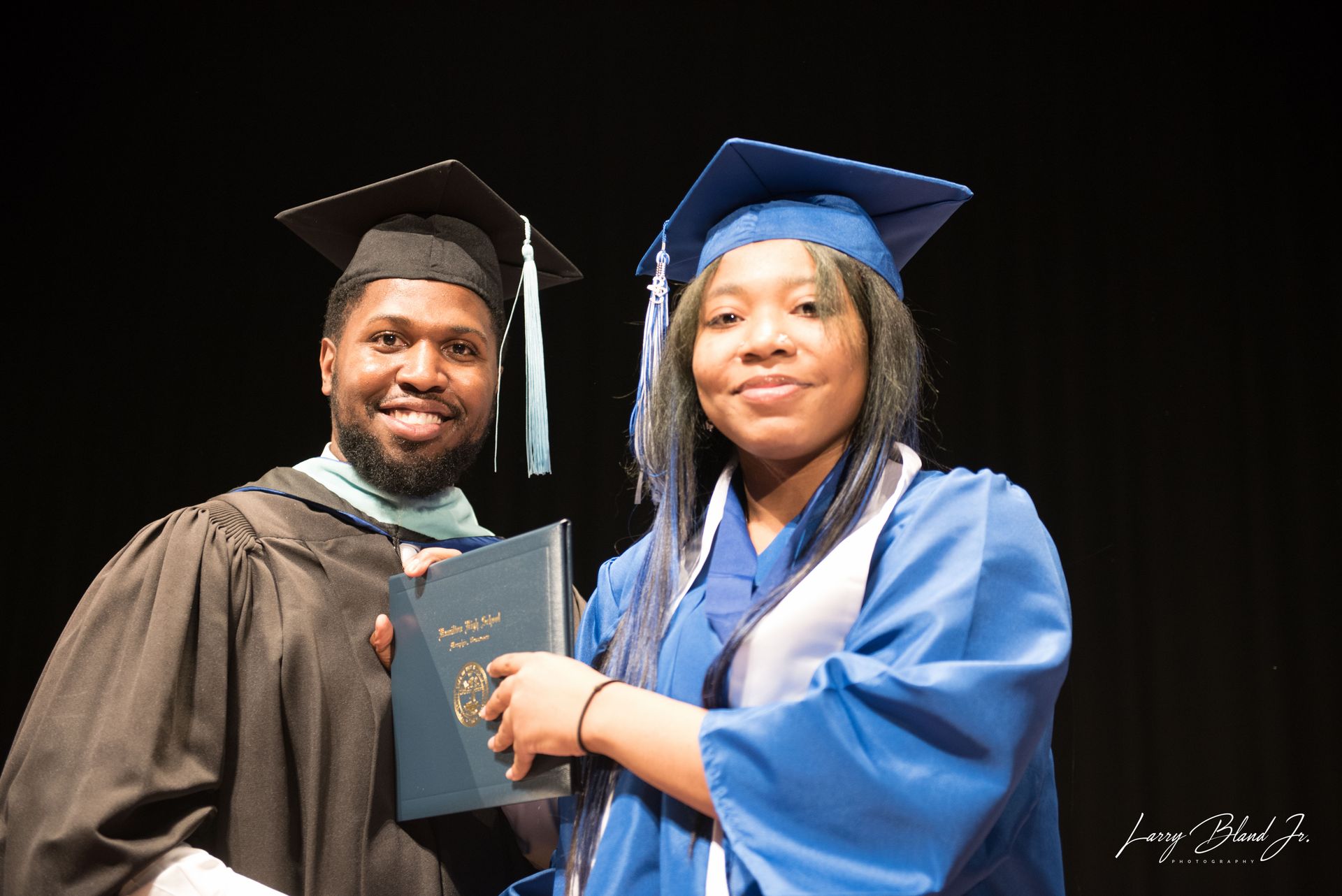 Man in black robe hands diploma to person in blue cap and gown.