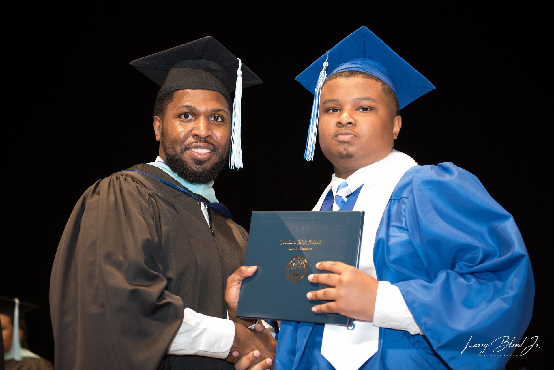 Two people in graduation attire shake hands. One presents a diploma, both smiling on stage.