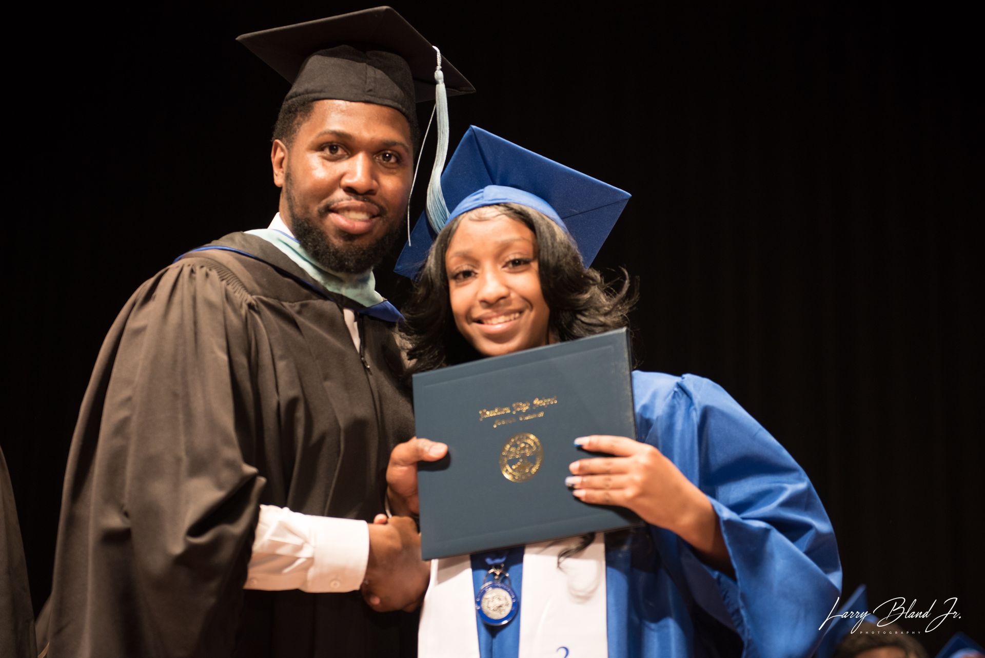 Person in graduation gown hands diploma to person in blue cap and gown.