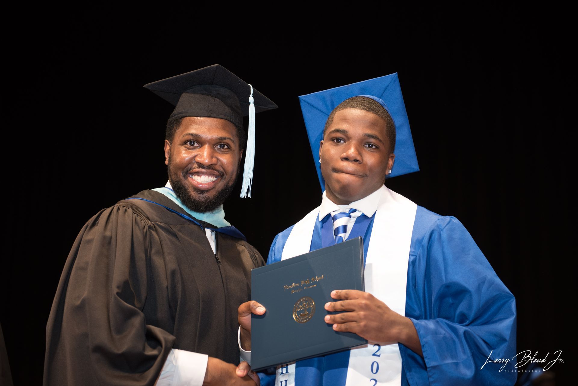 Man in black graduation gown shakes hands with graduate in blue gown, holding diploma.