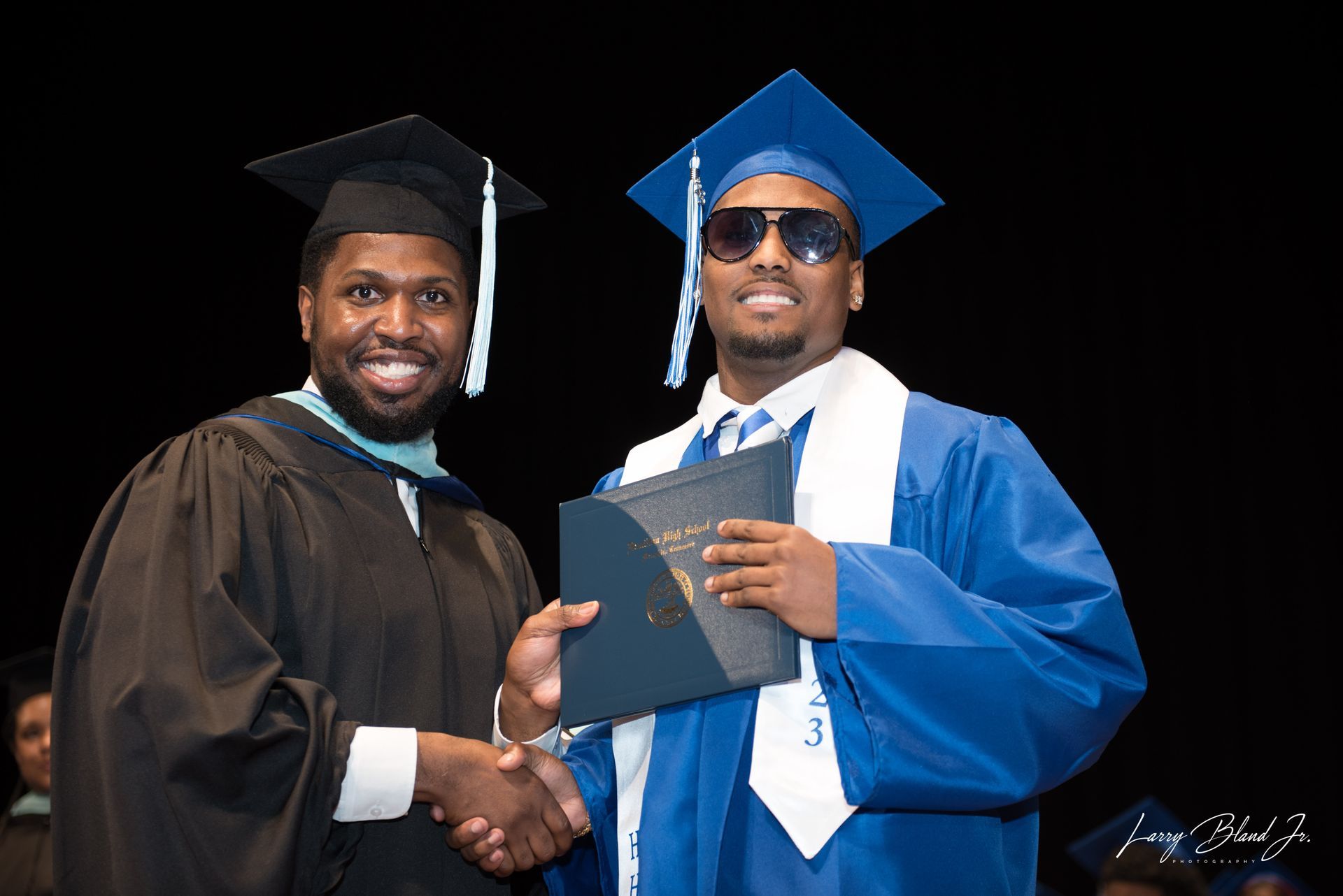 Man in blue graduation gown receives diploma, shakes hands with another in black gown.