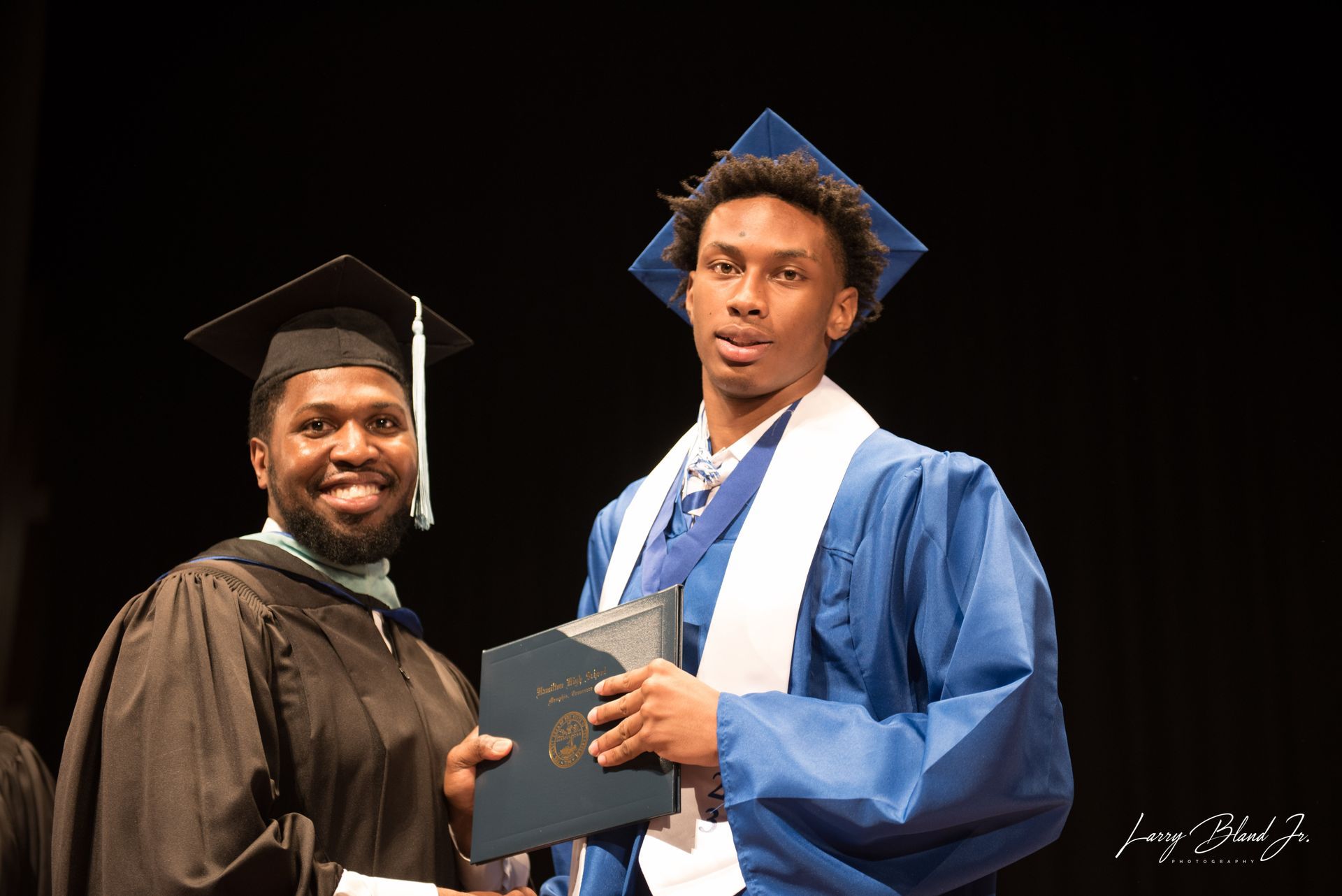 Man in black graduation gown presents diploma to a person in blue cap and gown.