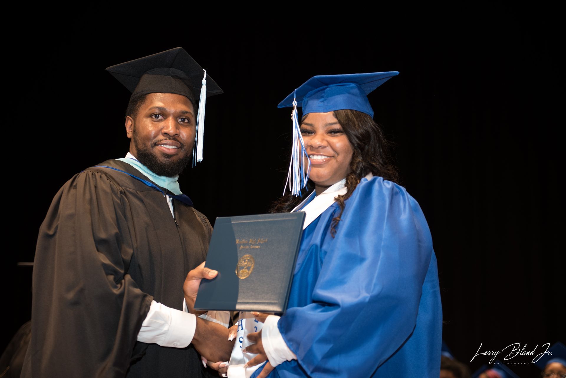 Man in black robe hands diploma to woman in blue robe on stage.