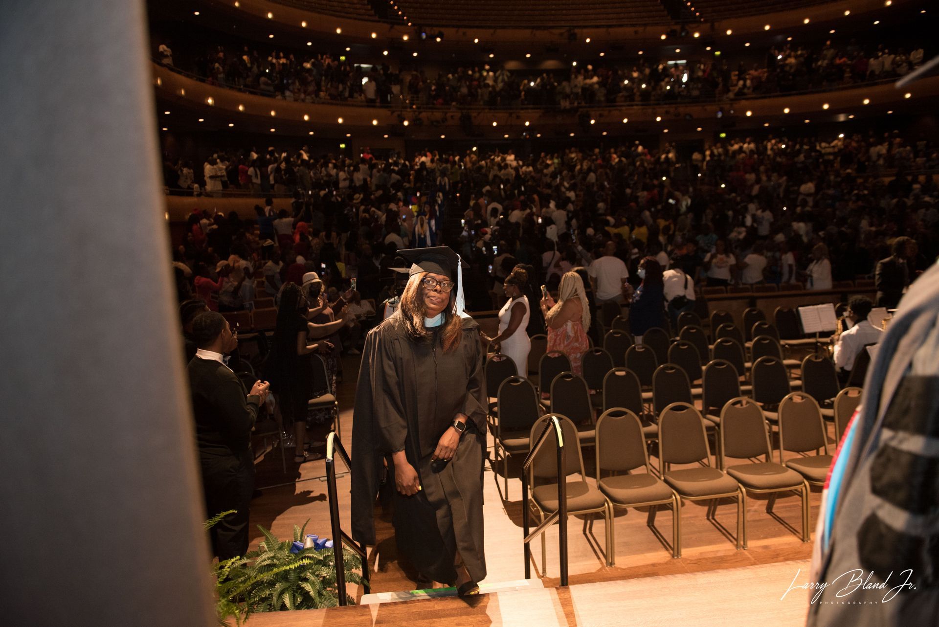 Graduate in cap and gown walks off stage in a crowded auditorium; audience applauds.