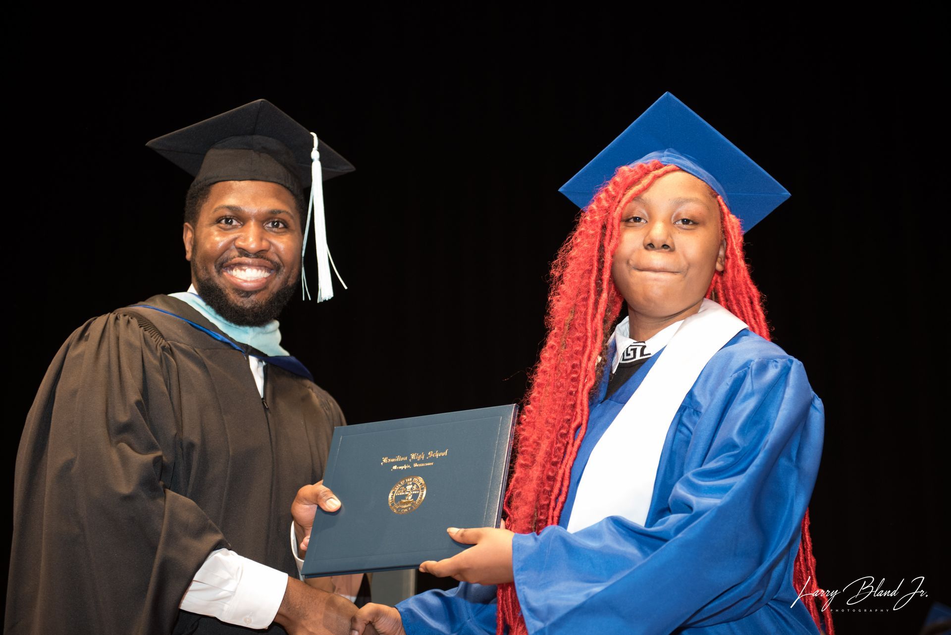Person in graduation gown presents diploma to graduate in blue gown.