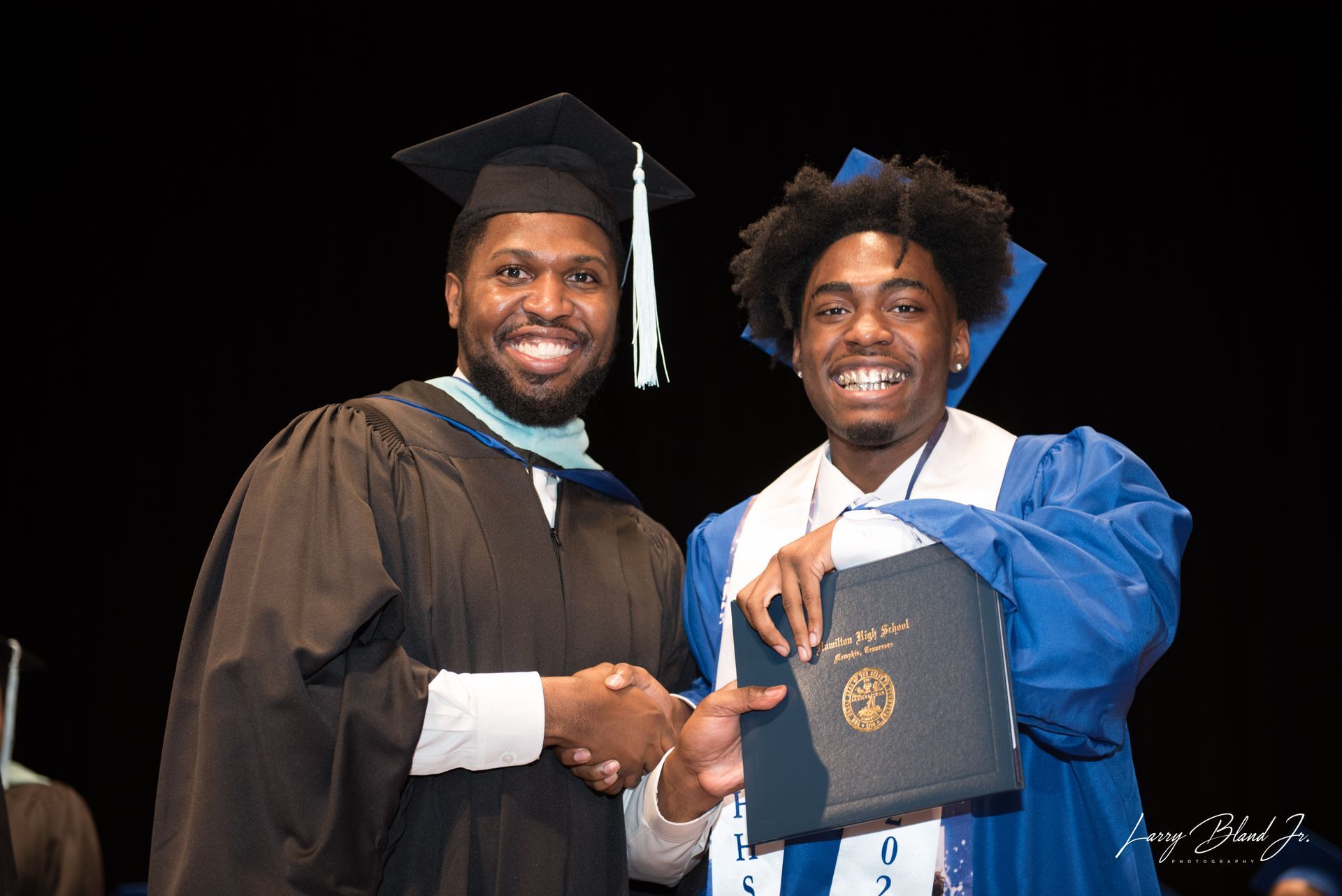 Man in cap and gown shakes hands with a graduate in blue gown, receiving a diploma.