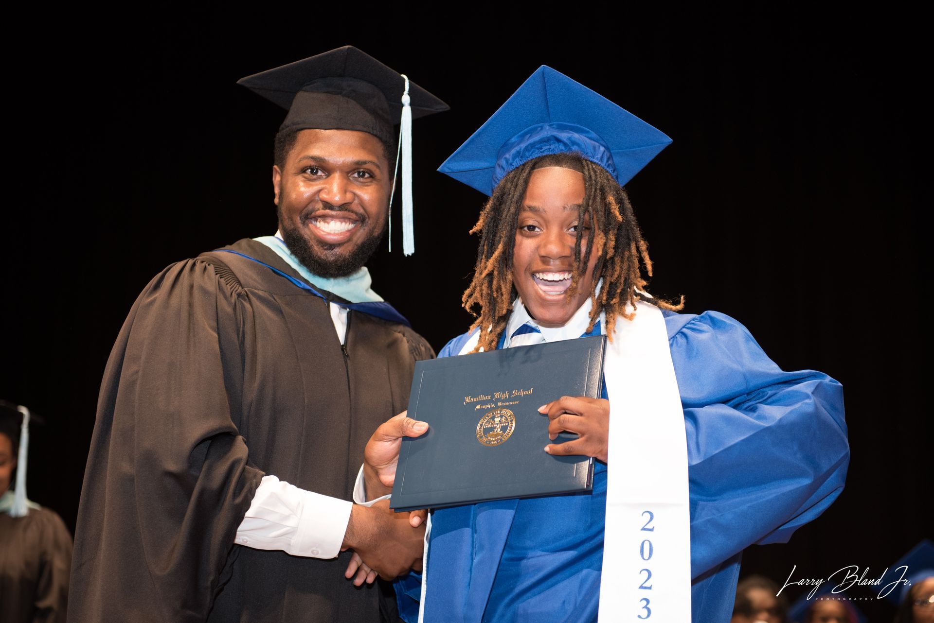 Man in cap and gown handing diploma to graduate in blue cap and gown on stage.
