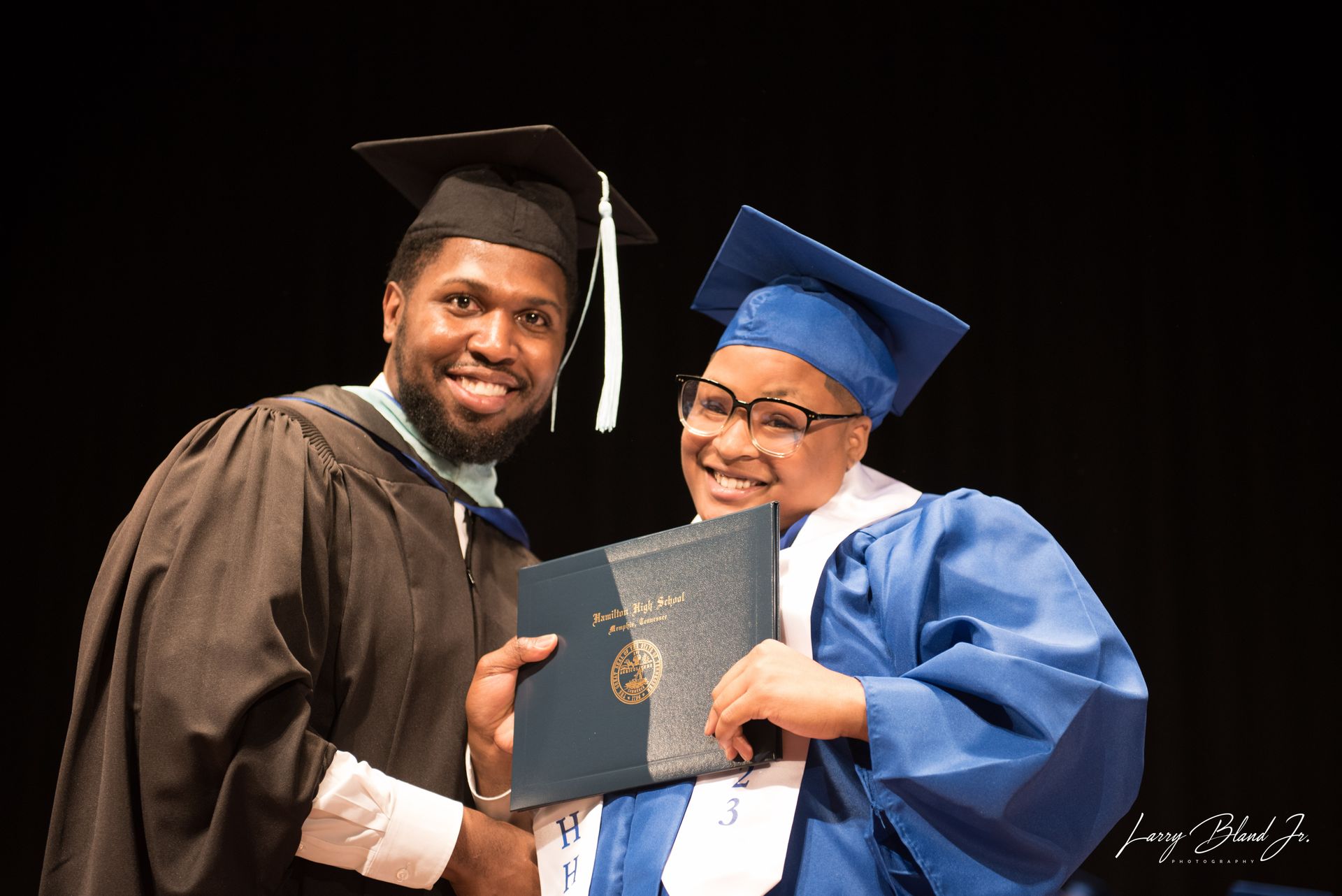 Two graduates in caps and gowns smiling, one holding a diploma, black backdrop.