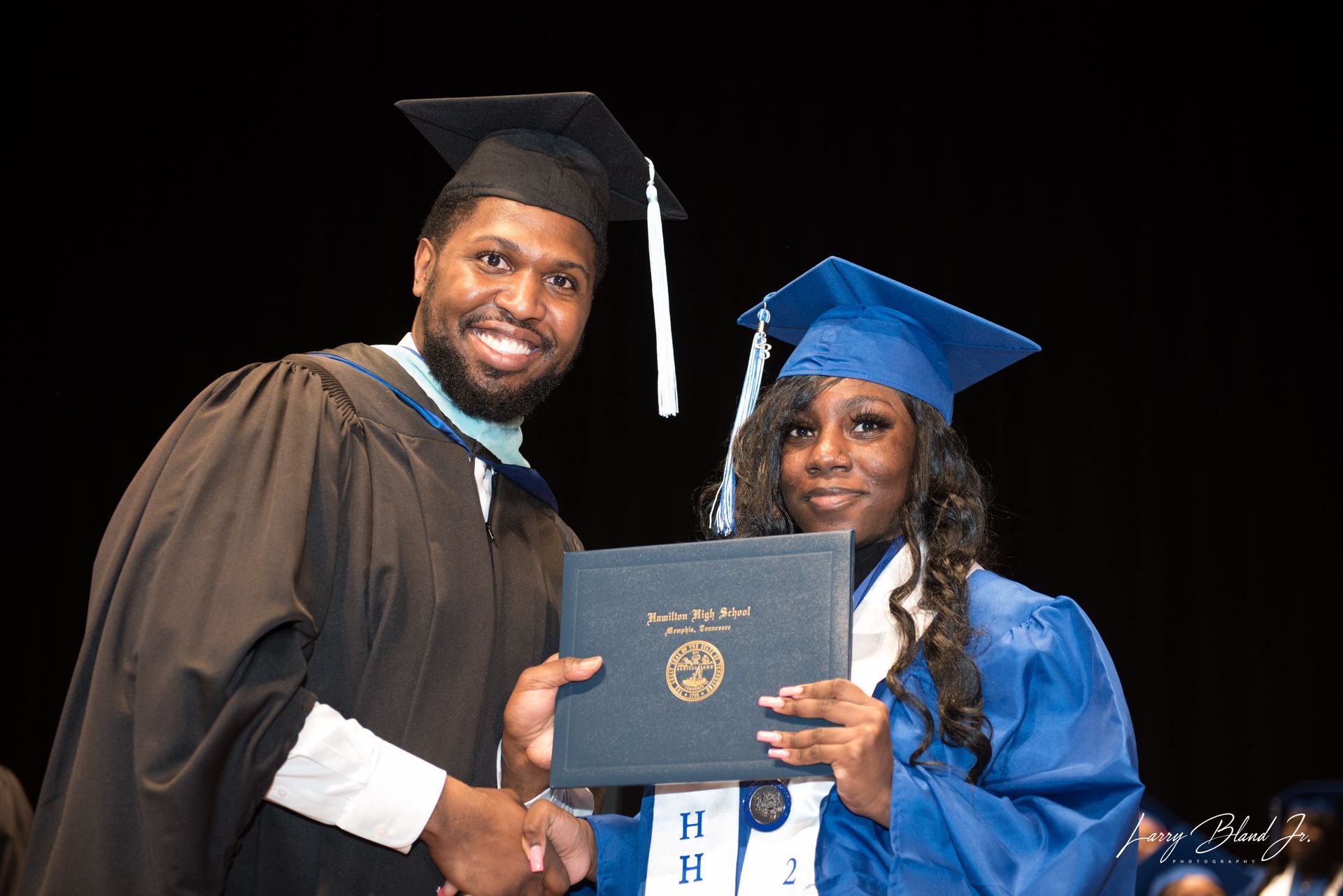 Person handing a diploma to a graduate wearing a blue cap and gown. Both are smiling at a graduation ceremony.