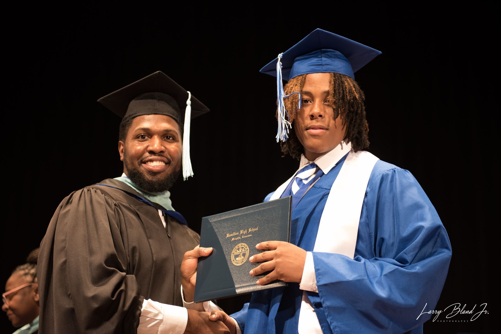 Graduate receiving diploma at ceremony; blue and black gowns, handshake.