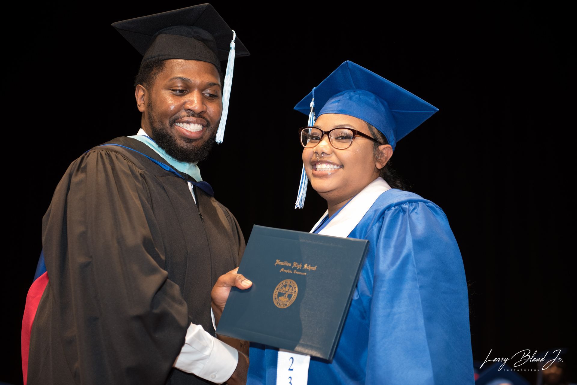 Person in black graduation gown hands diploma to a person in blue gown; stage backdrop.