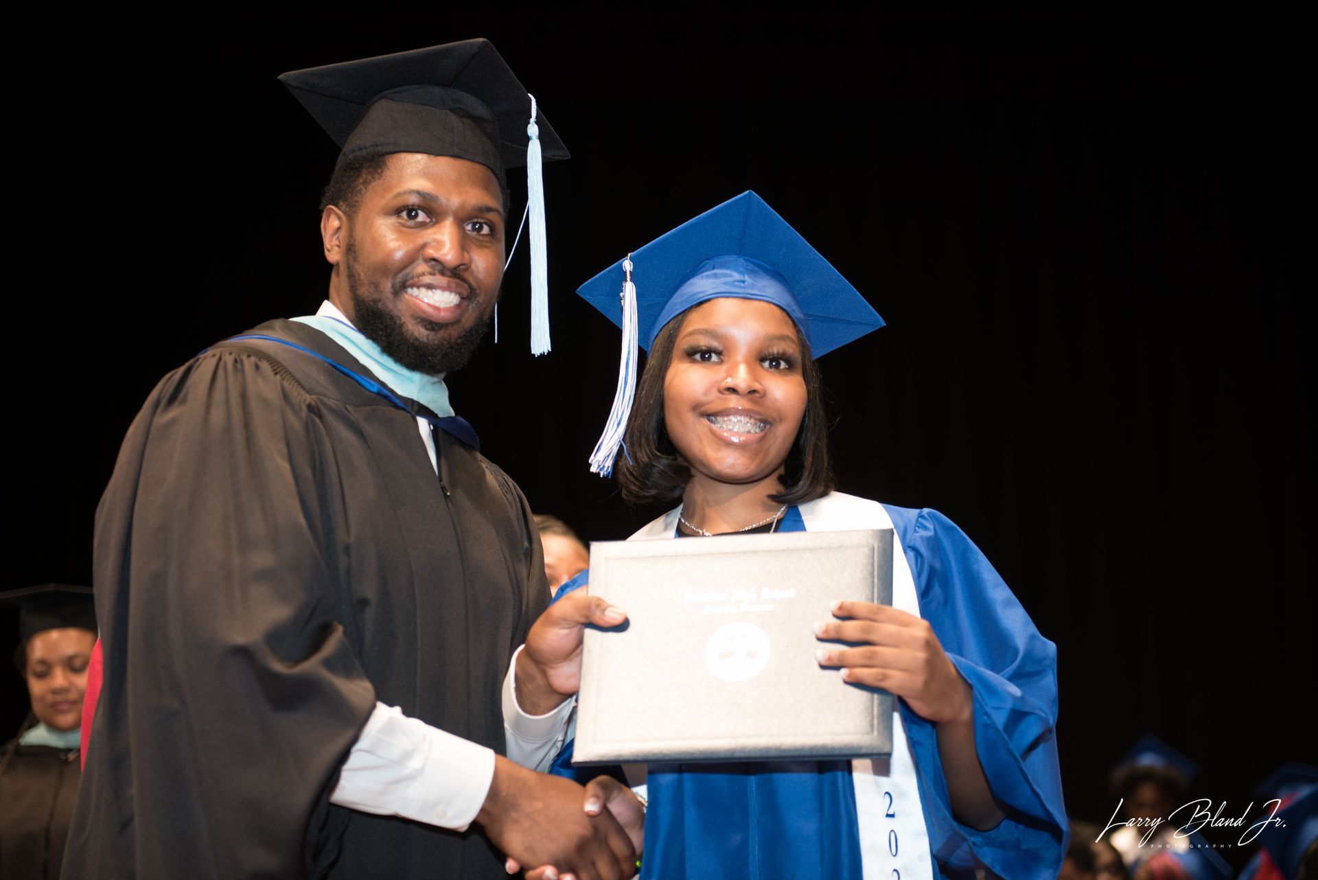 Man in graduation gown hands diploma to person in blue cap and gown on stage.